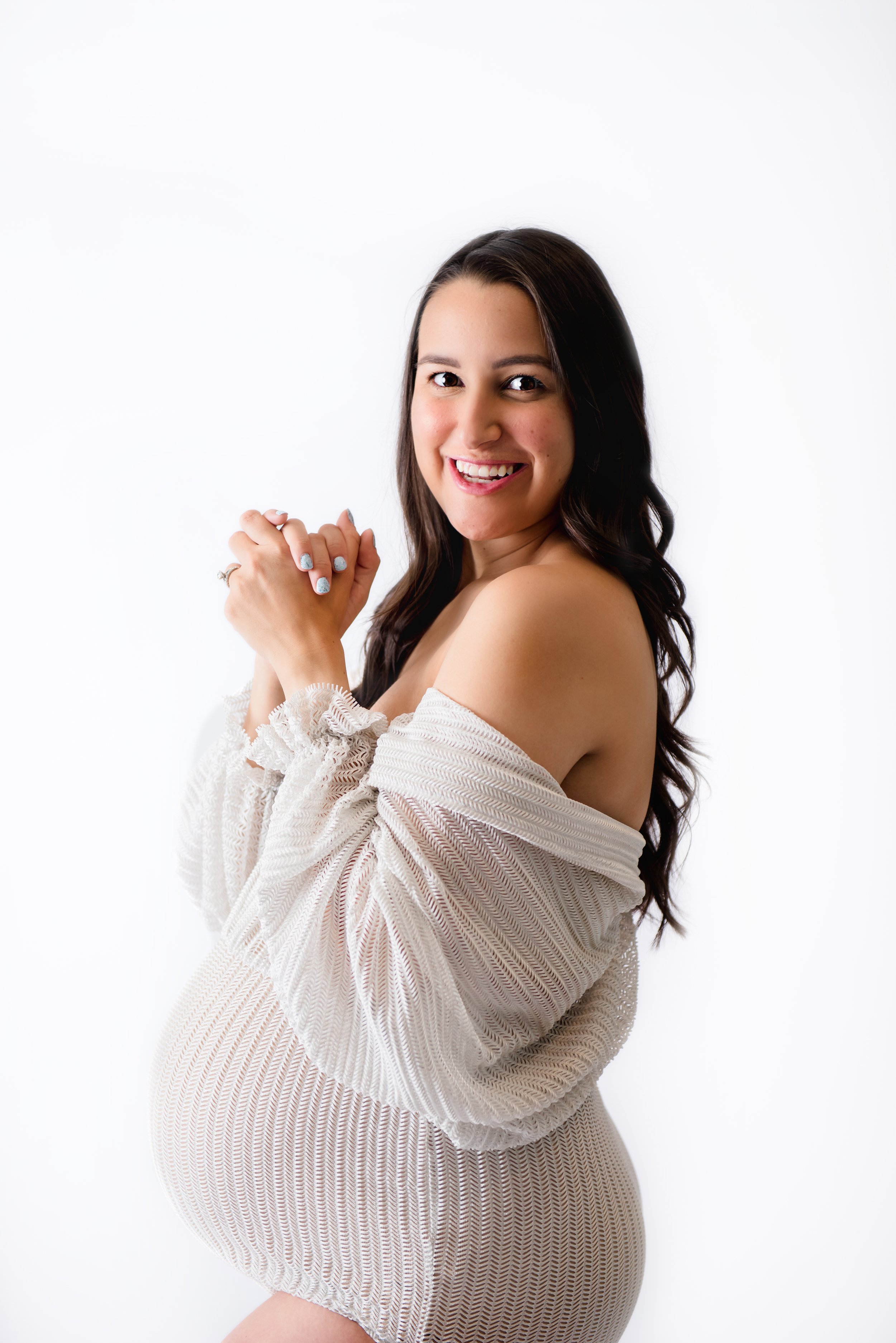 Fine art maternity portrait of a smiling mother-to-be in an off-the-shoulder beige knit dress, photographed against a clean white studio background for a luxury portrait experience near Stone Oak.