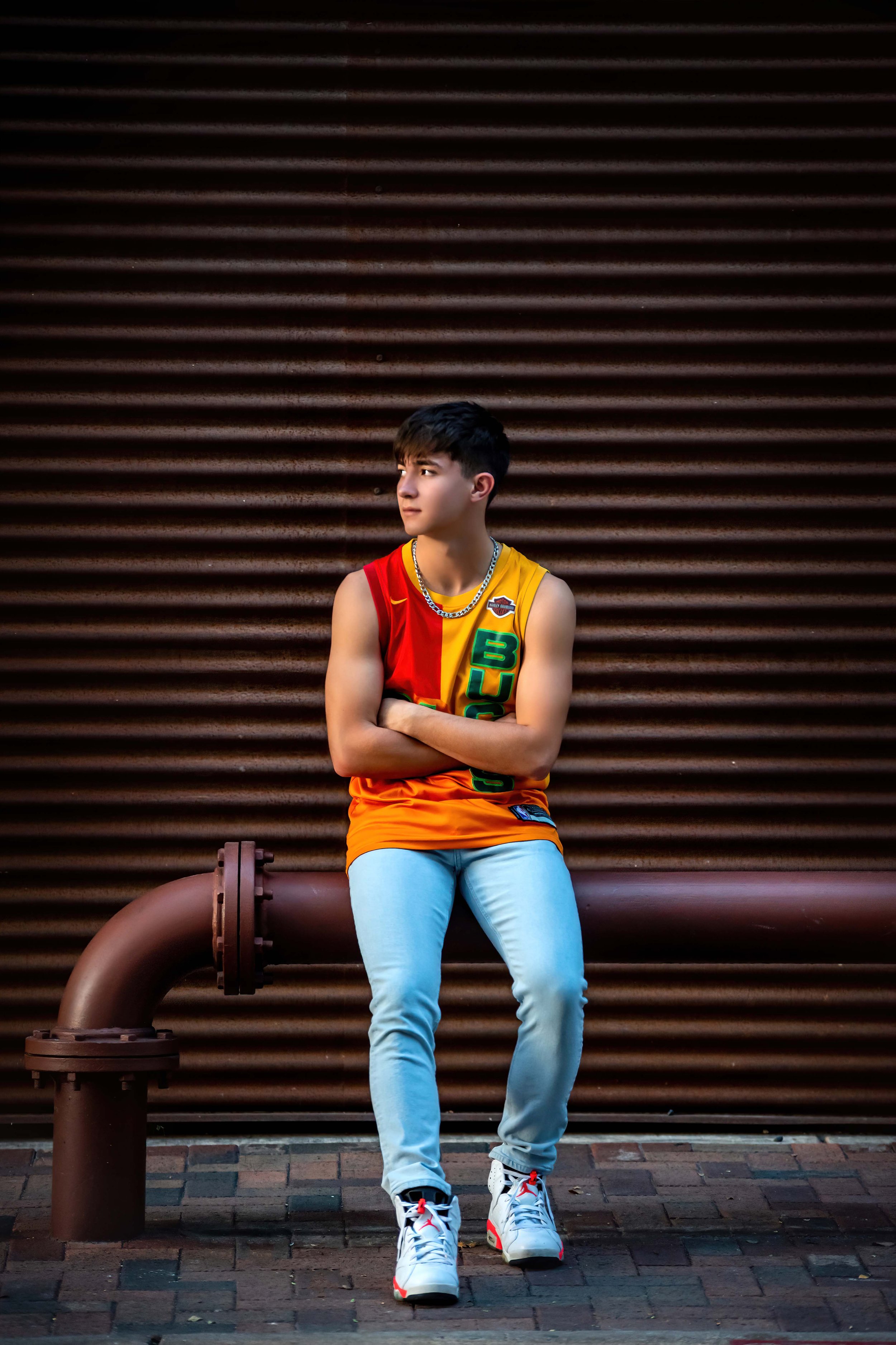 A young man with dark hair wearing a colorful sports jersey, light jeans, and white sneakers is leaning with his back against a large rusty pipe and a brown corrugated metal wall in an urban setting.