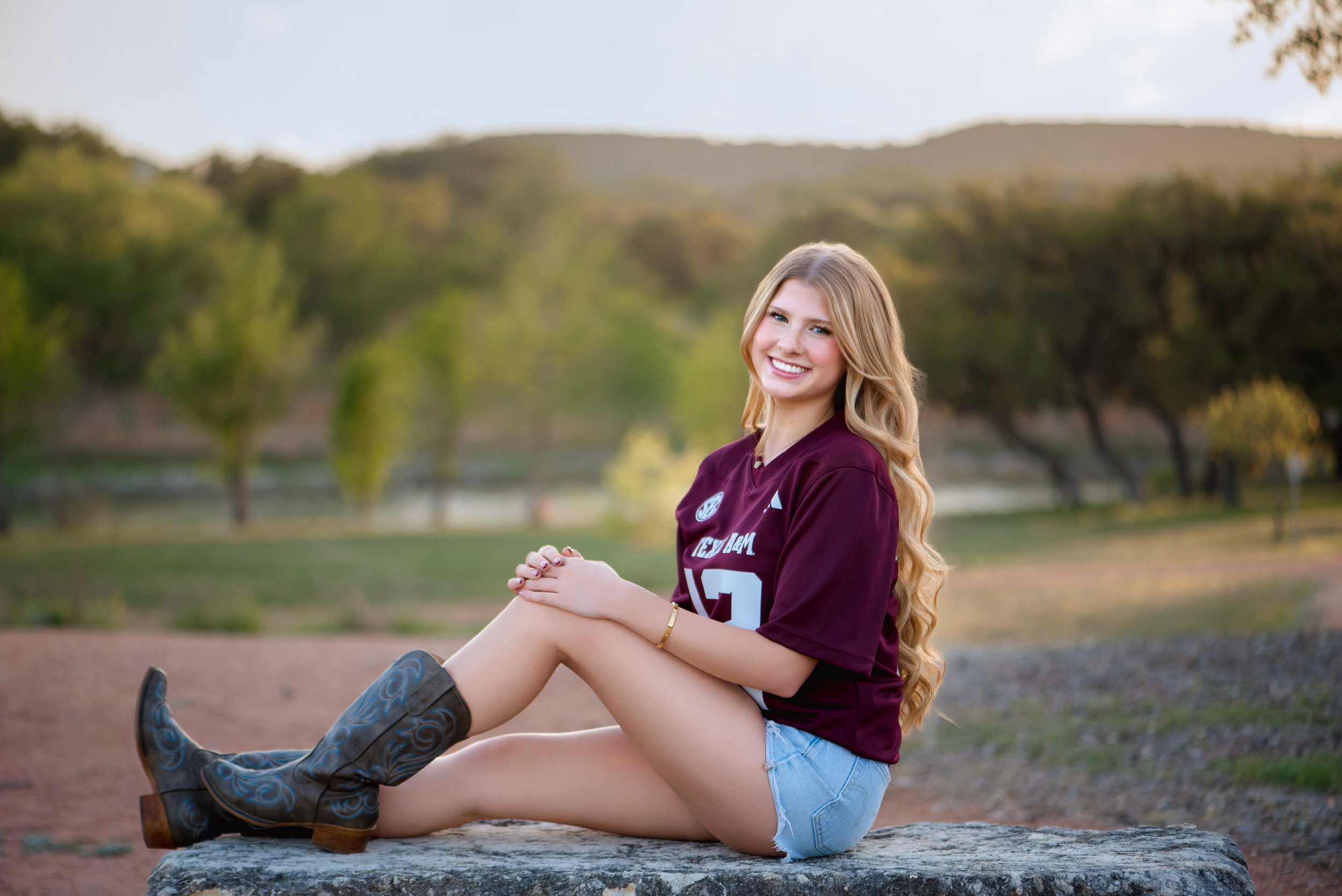 Young woman with long blond hair sitting on a rock outdoors, wearing a maroon sports jersey, denim shorts, and cowboy boots, smiling at the camera with a scenic natural background.