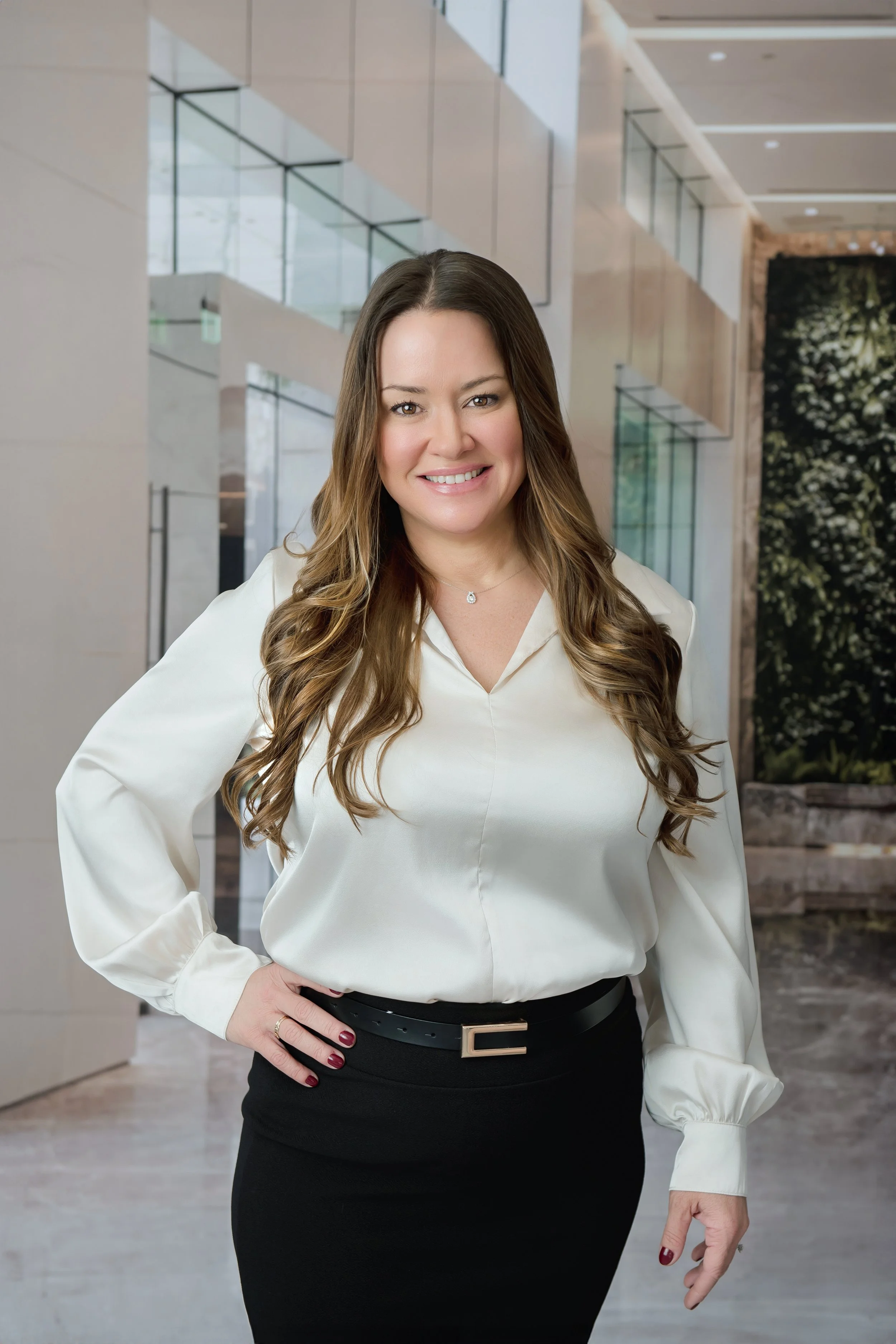 A woman with long, wavy brown hair smiling and standing in a modern building lobby with glass walls and indoor greenery.