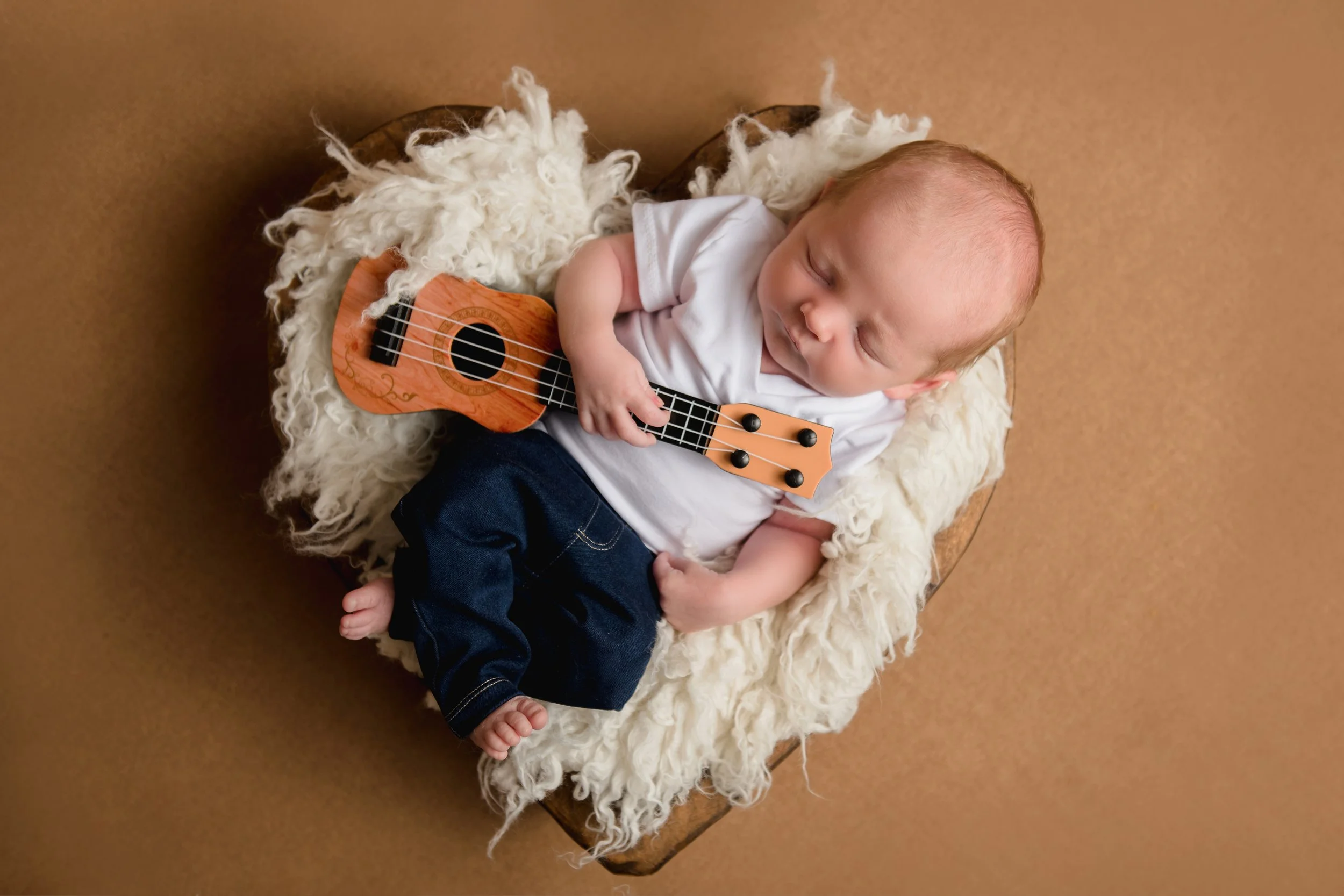 A sleeping baby lying on a fluffy white blanket in a basket, holding a small ukulele.