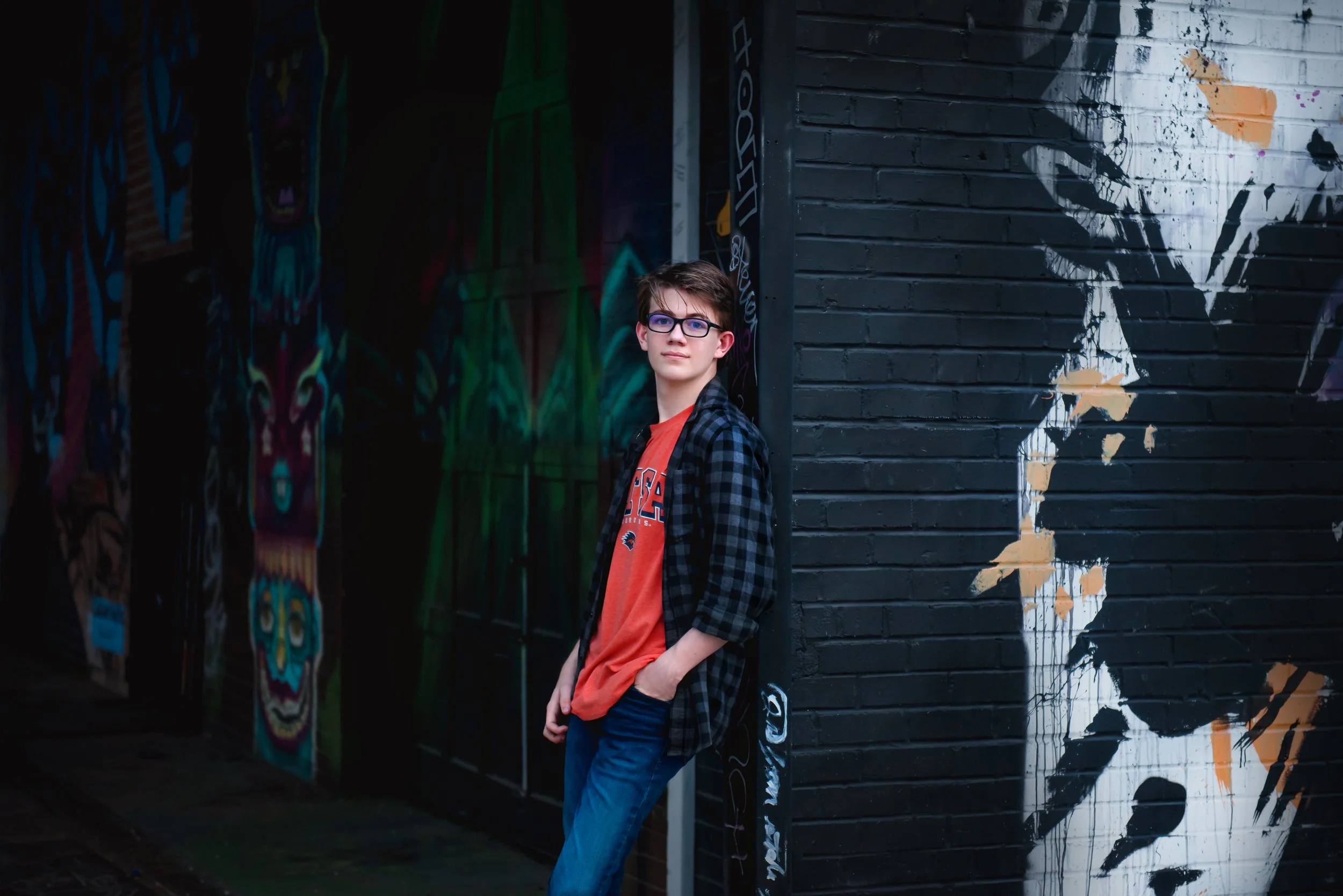A young man with glasses, wearing a black and blue checkered shirt over an orange T-shirt, stands against a black brick wall featuring graffiti art, including a large stylized face and a painted animal figure on a nearby wall.