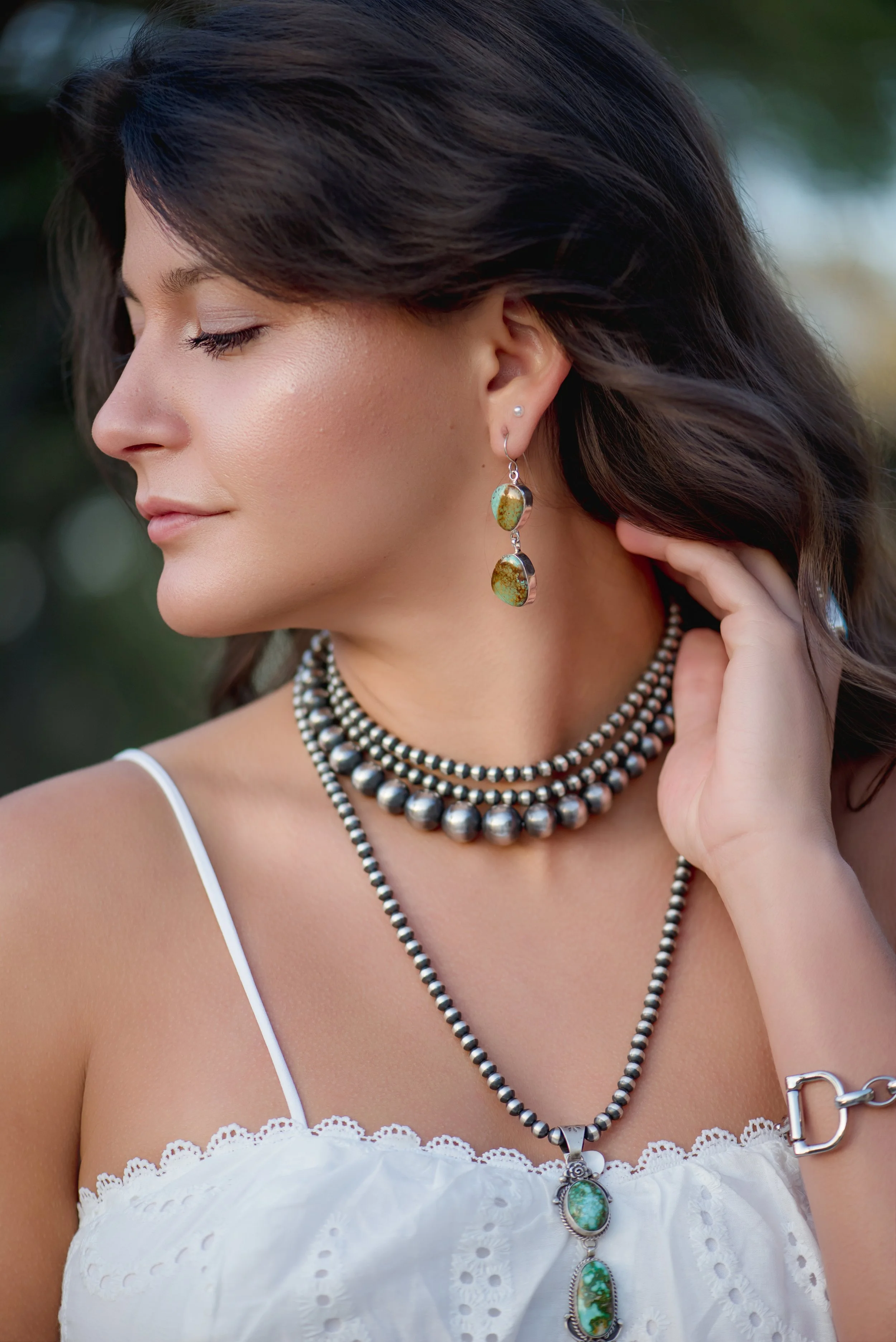 A woman with dark hair wearing layered silver jewelry, including beaded necklaces, earrings with stones, and a bracelet, dressed in a white top with lace details, outdoors with blurred green background.