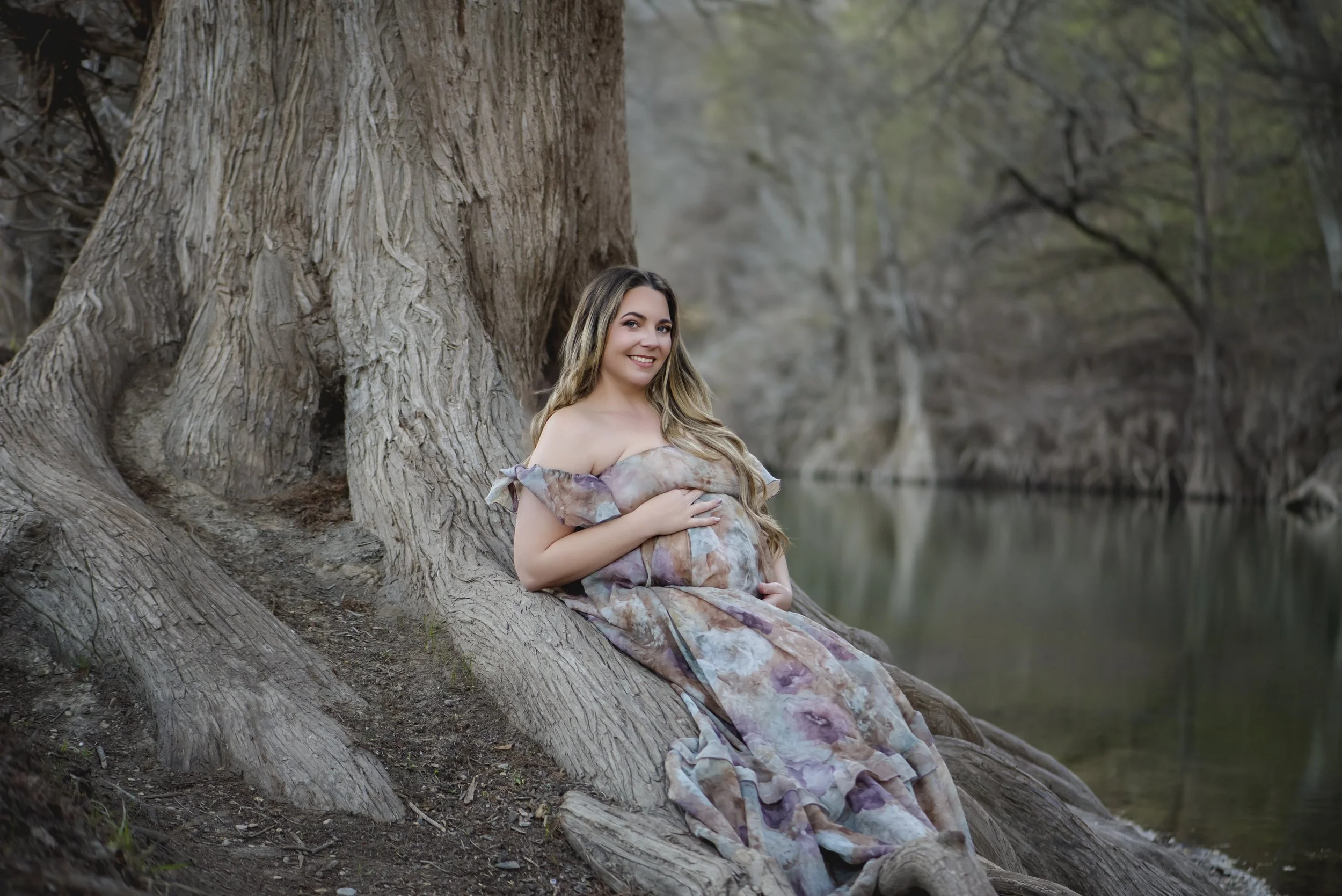 Fine art portrait of a young woman in a floral dress seated on a tree root beside a river in the Texas Hill Country near Spring Branch.