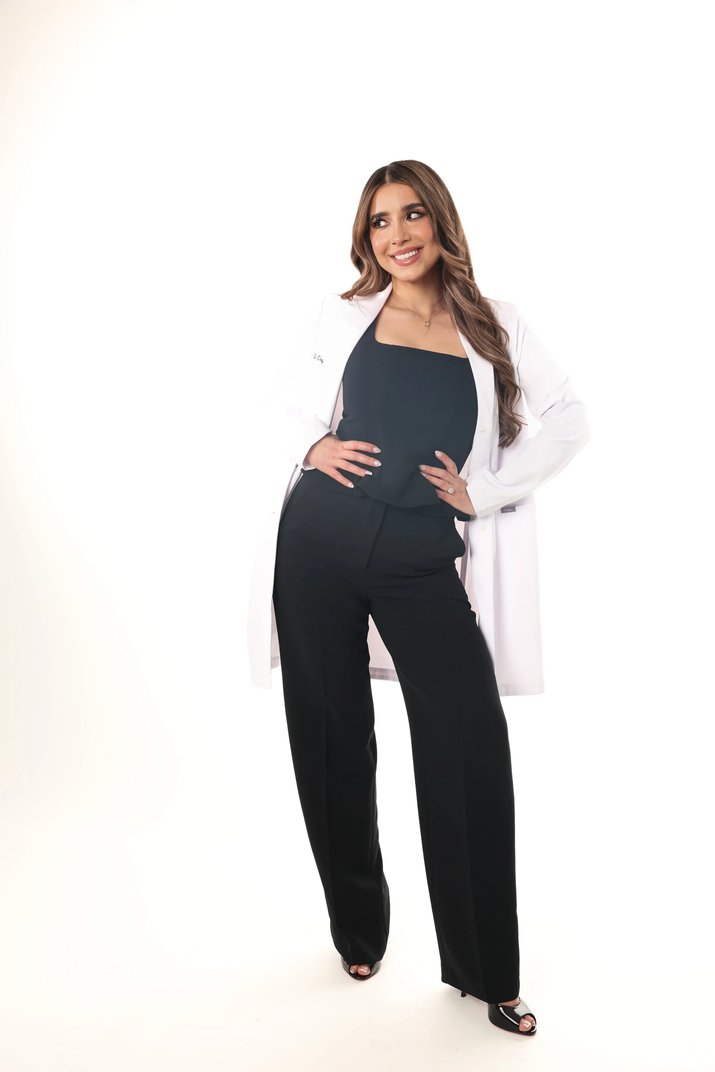 A woman in professional attire, wearing a white lab coat, black top, and black pants, standing with her hands on her hips and smiling.