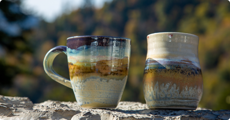 Coffee mugs featuring Smoky Mountain Autumn glaze are set on a rock outside with the Smoky Mountains in the distance