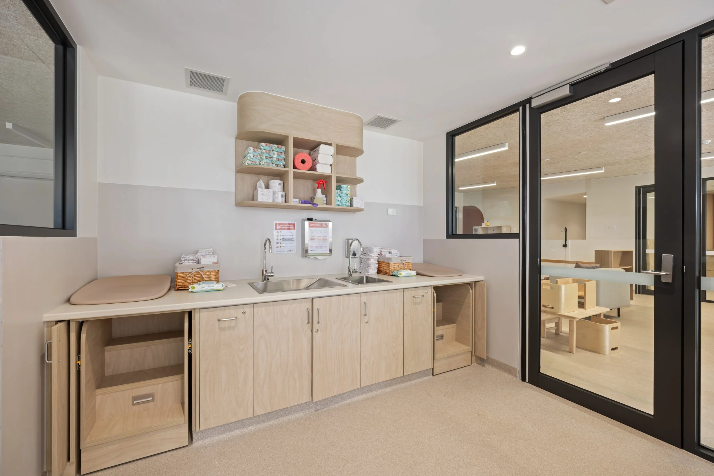 A clean, modern nursing station with a light wood countertop and cabinets, dual sinks, and storage shelves, adjacent to a glass sliding door leading to a pediatric or daycare play area with wooden furniture.