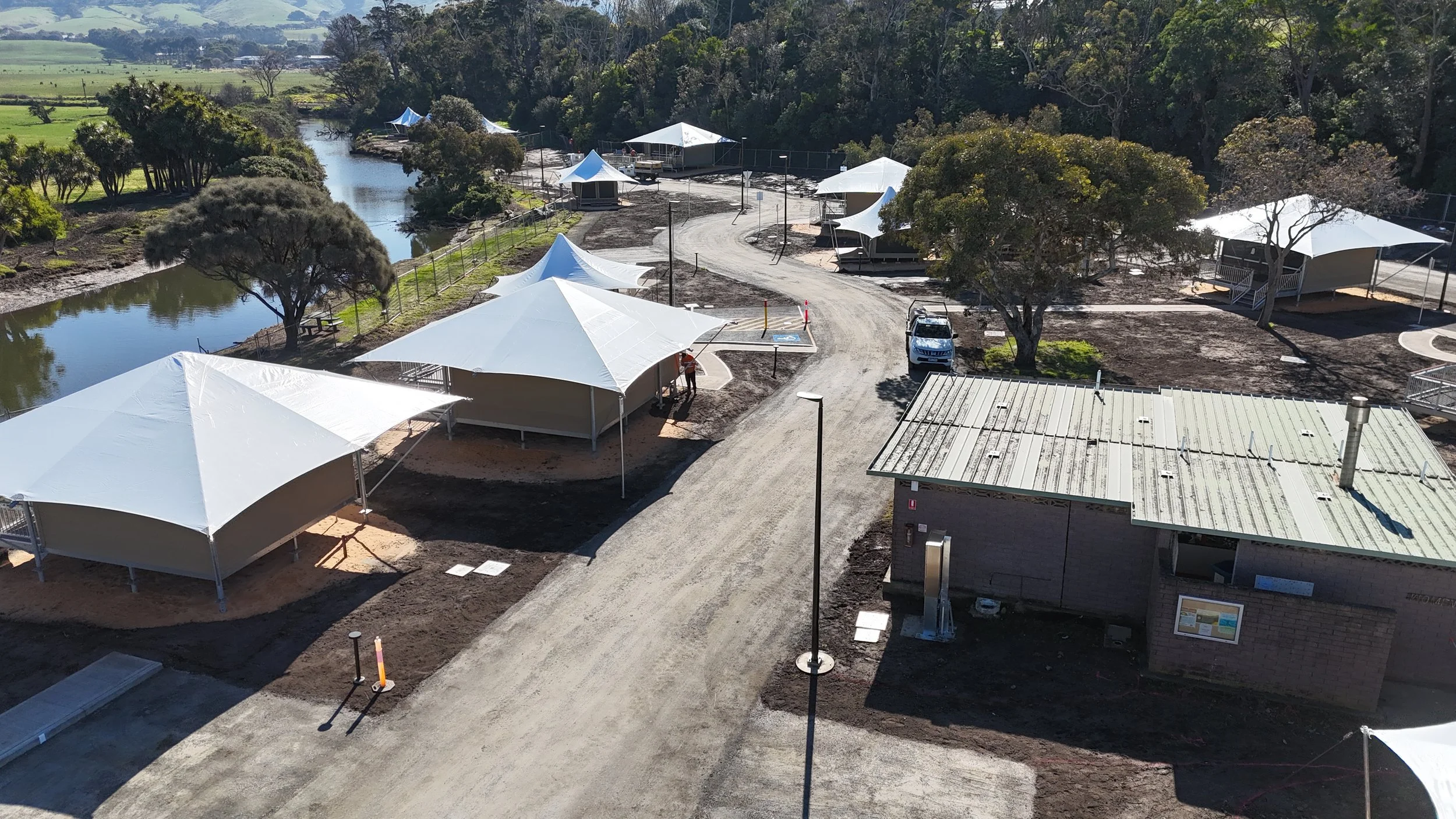 Aerial view of a construction site near a river, with multiple tents and a parked vehicle, surrounded by trees and open fields.