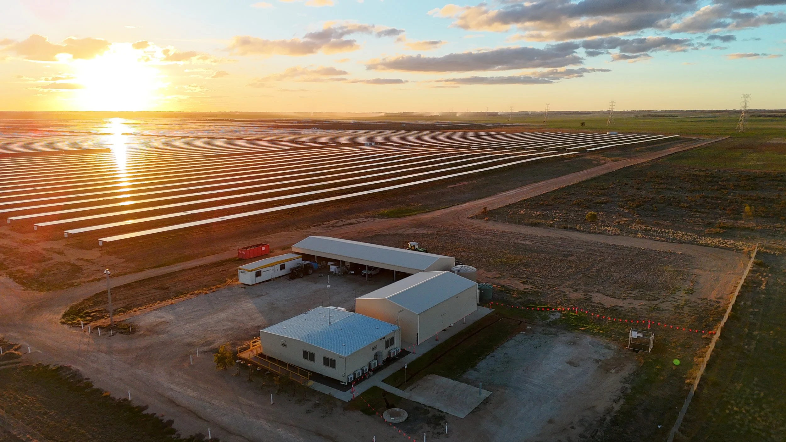 A solar farm with numerous solar panels extending across the land at sunset, with a few small buildings and dirt roads in the foreground.