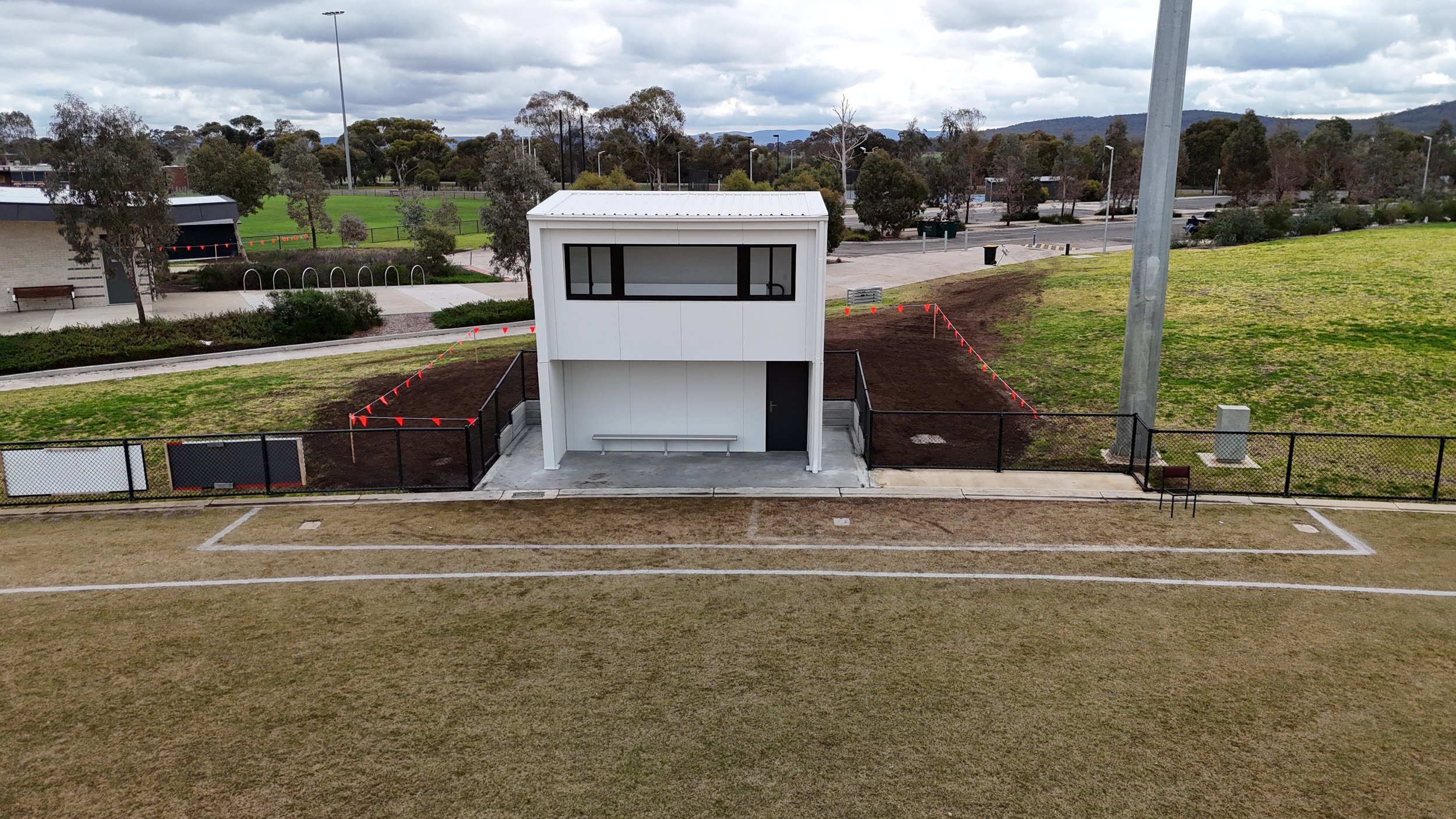 A small white building with large windows, located in a park or sports field, surrounded by a black fence. The field in the foreground is brown with white lines, possibly a running track. In the background, there are trees, parked cars, and a cloudy sky.