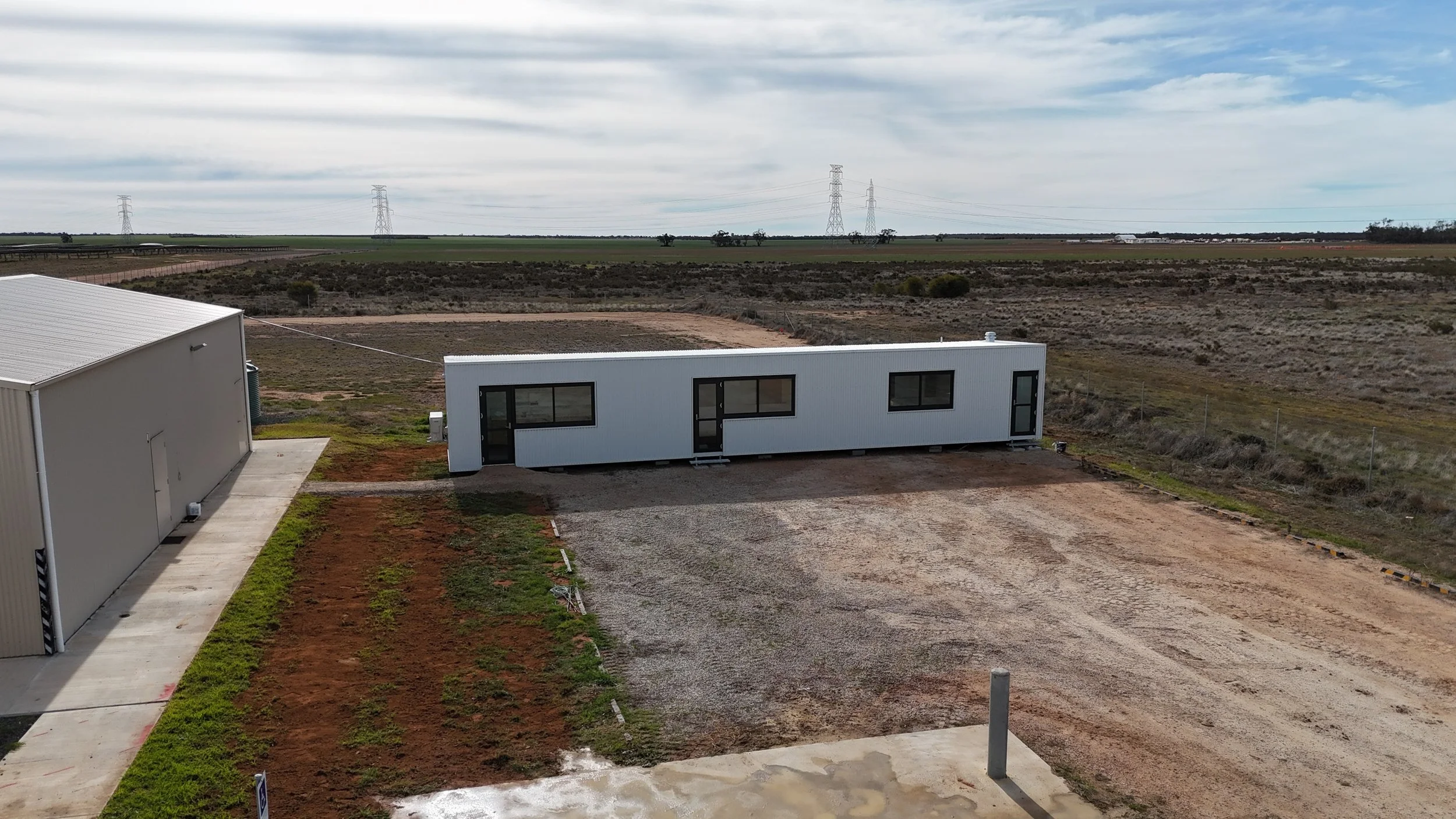 A landscape with two white modular buildings and a gravel driveway in the foreground, open fields, power lines, and a cloudy sky in the background.