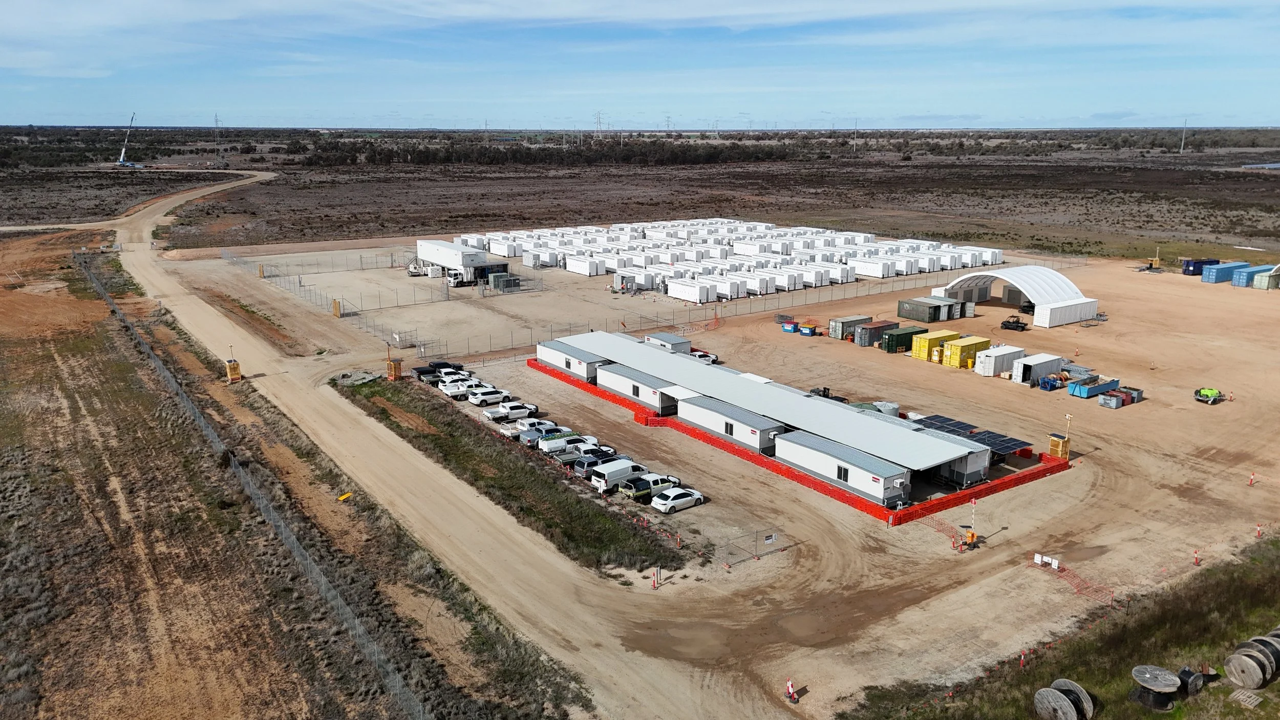 An industrial site with numerous white storage containers, trucks, and multiple temporary buildings in a remote, arid landscape with dirt roads and sparse vegetation.