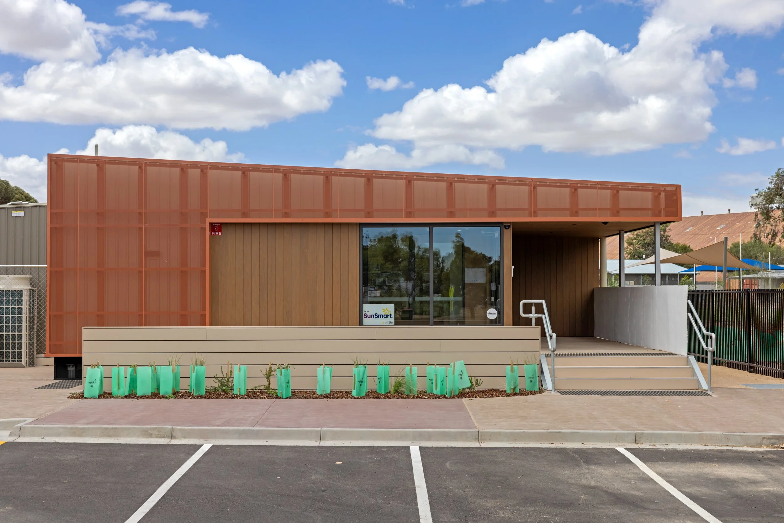 A modern building with wood paneling and a large glass door, with parking spaces in front and green plants in the foreground. The sky is partly cloudy.