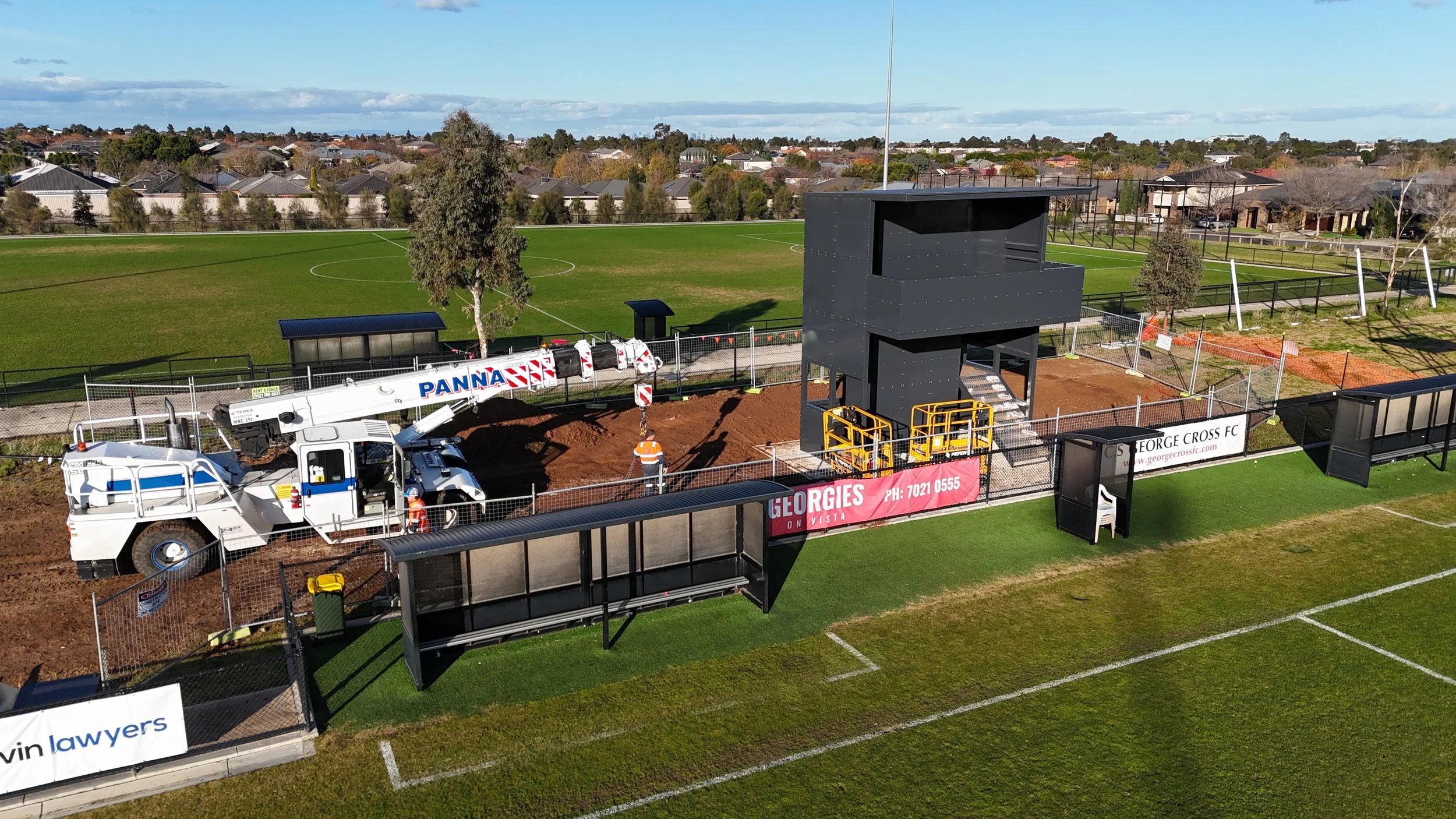 Construction site on a football field with a large black structure, construction vehicle, worker, fencing, and signage.