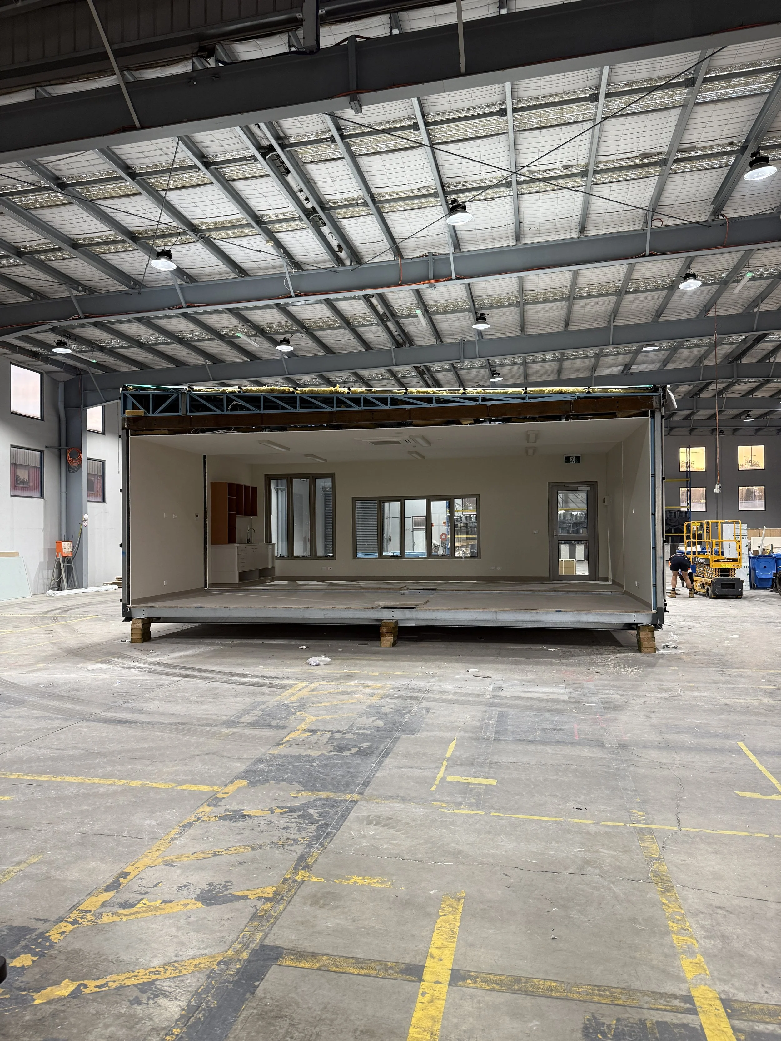 An interior view of a large industrial warehouse with a partially constructed prefabricated room or office unit in the center, supported by wooden blocks, with construction equipment and a worker in the background.
