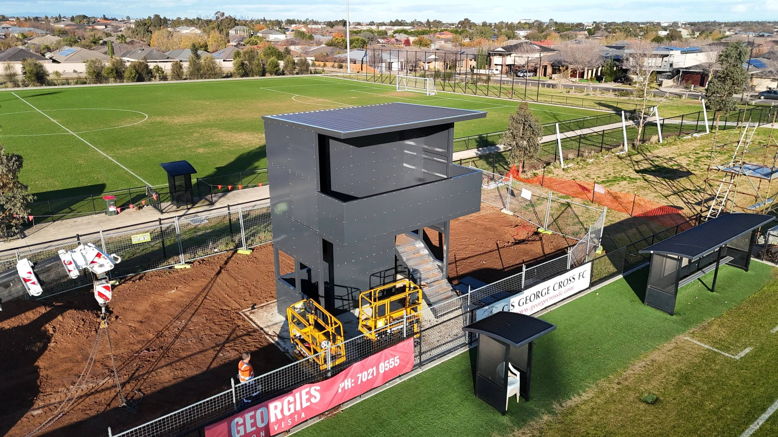 Construction site near a sports field with a gray building structure and a fence, with houses and trees in the background and clear blue sky.