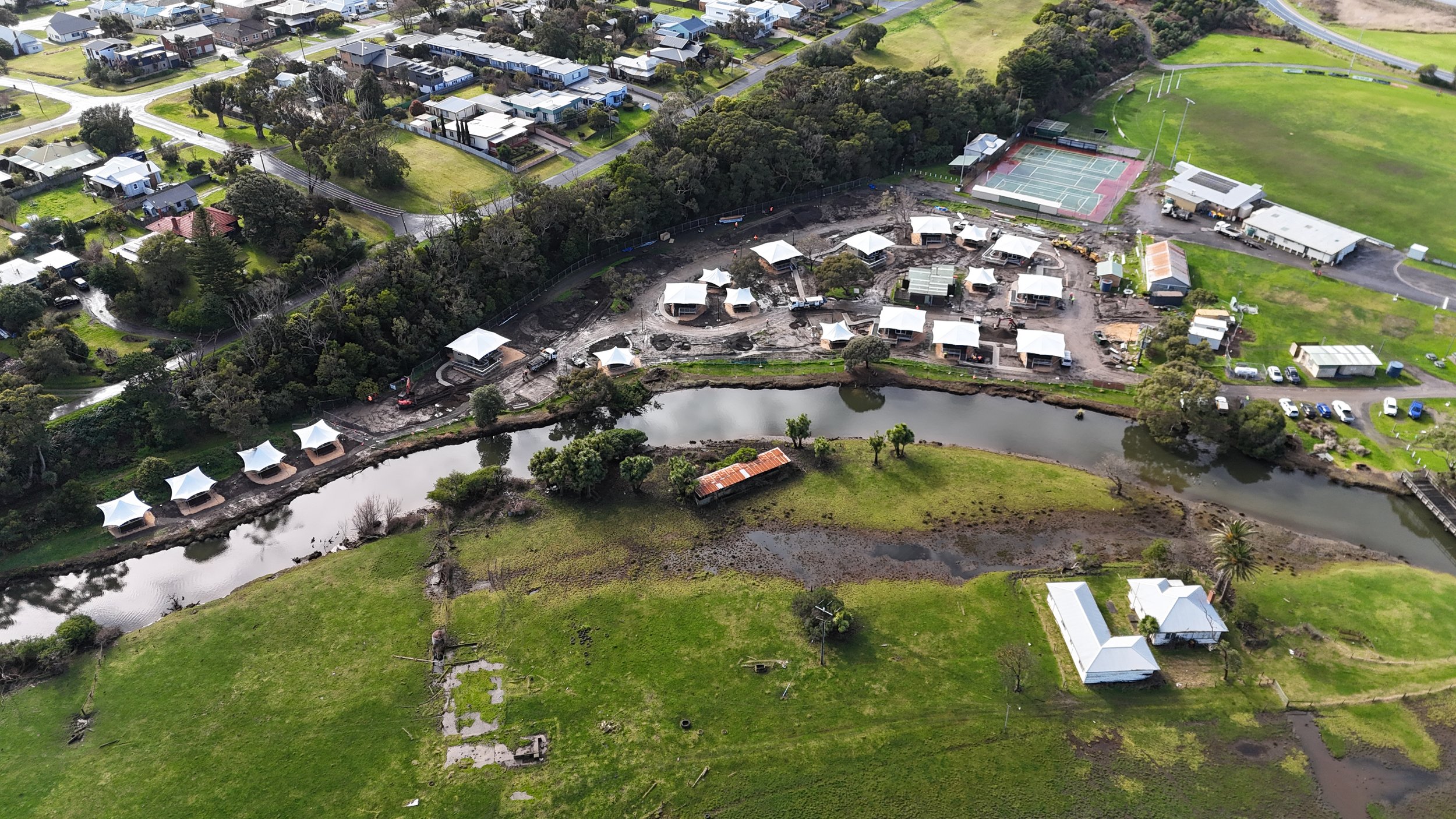 Aerial view of a construction site beside a river with houses, trees, and sports courts in the background.