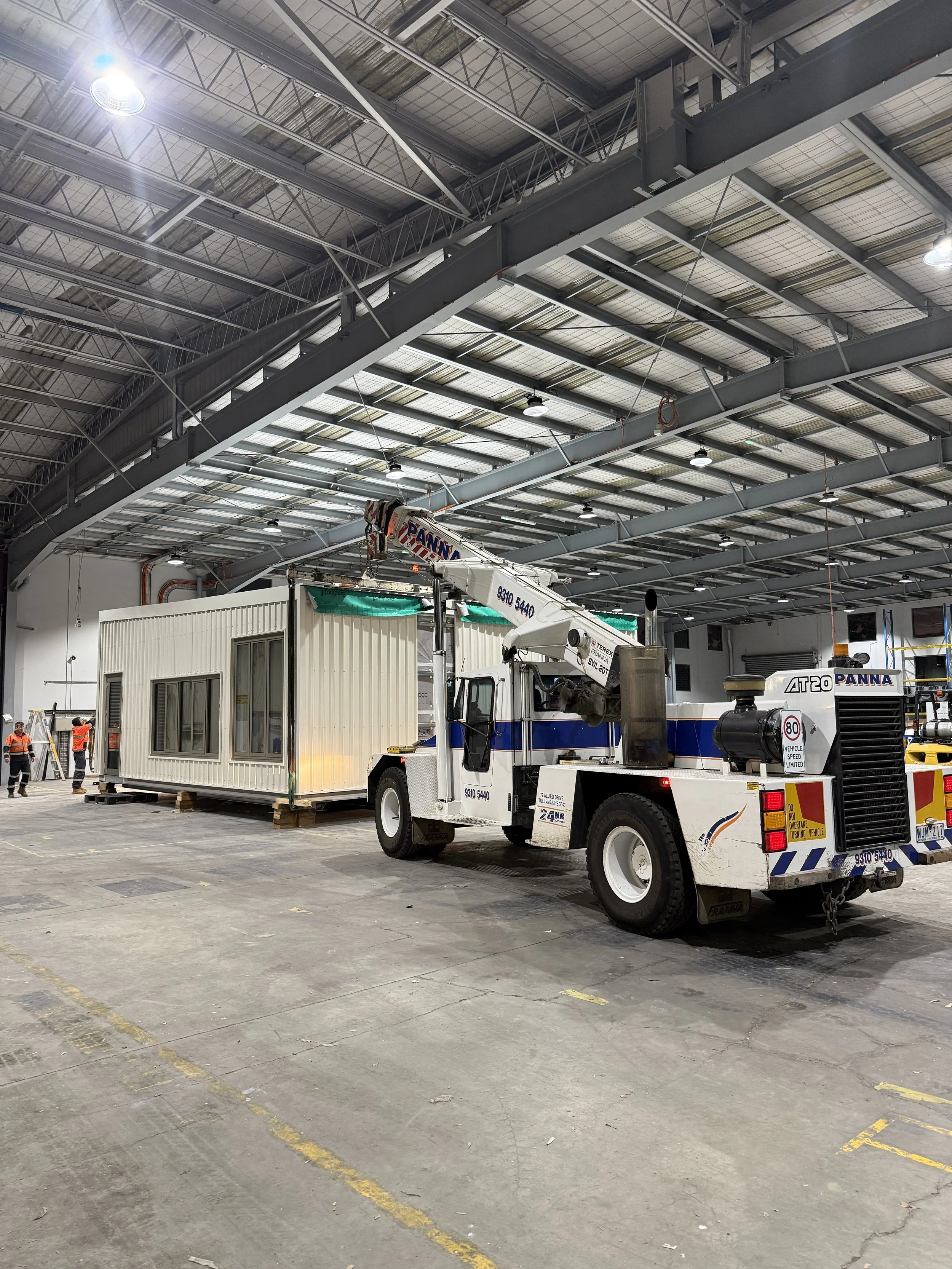 Inside a large industrial warehouse with a high ceiling, a white truck with a crane lifting a modular building unit. Several workers in orange safety vests are in the background.