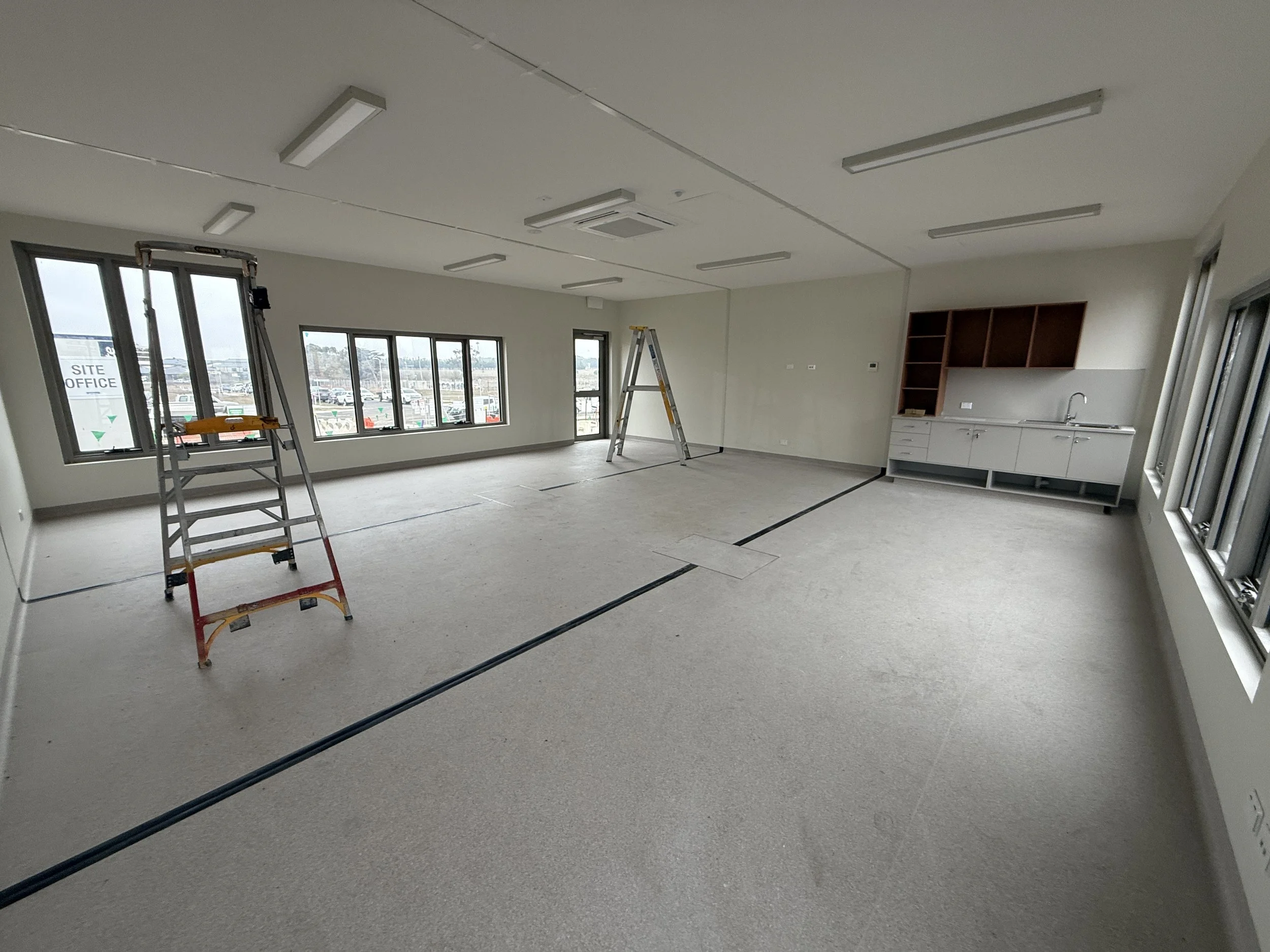 Empty room under construction with windows, ceiling lights, ladders, and a kitchen area.