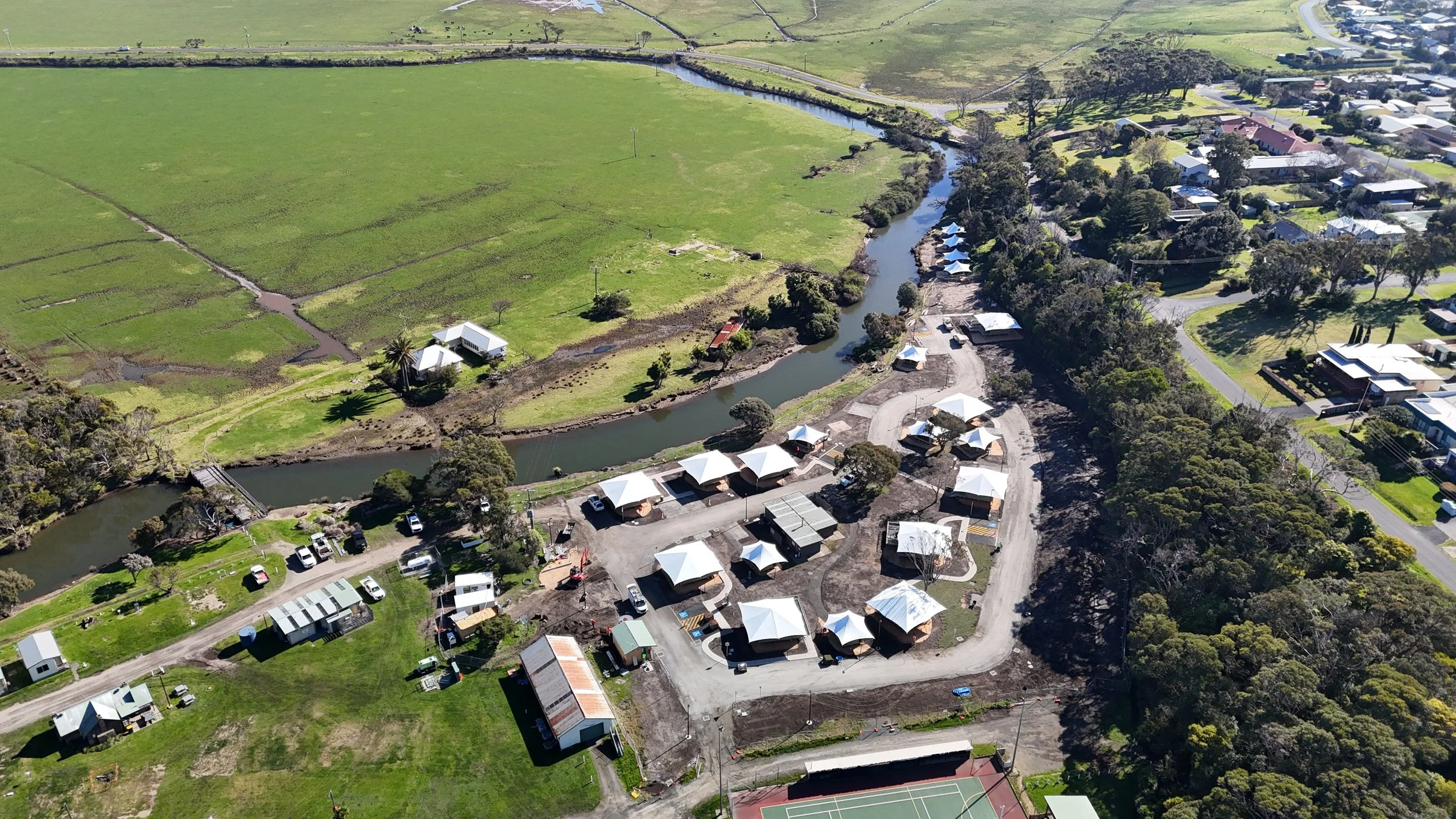 Aerial view of a park with a winding river, white tents, recreational areas, and surrounding residential neighborhood.