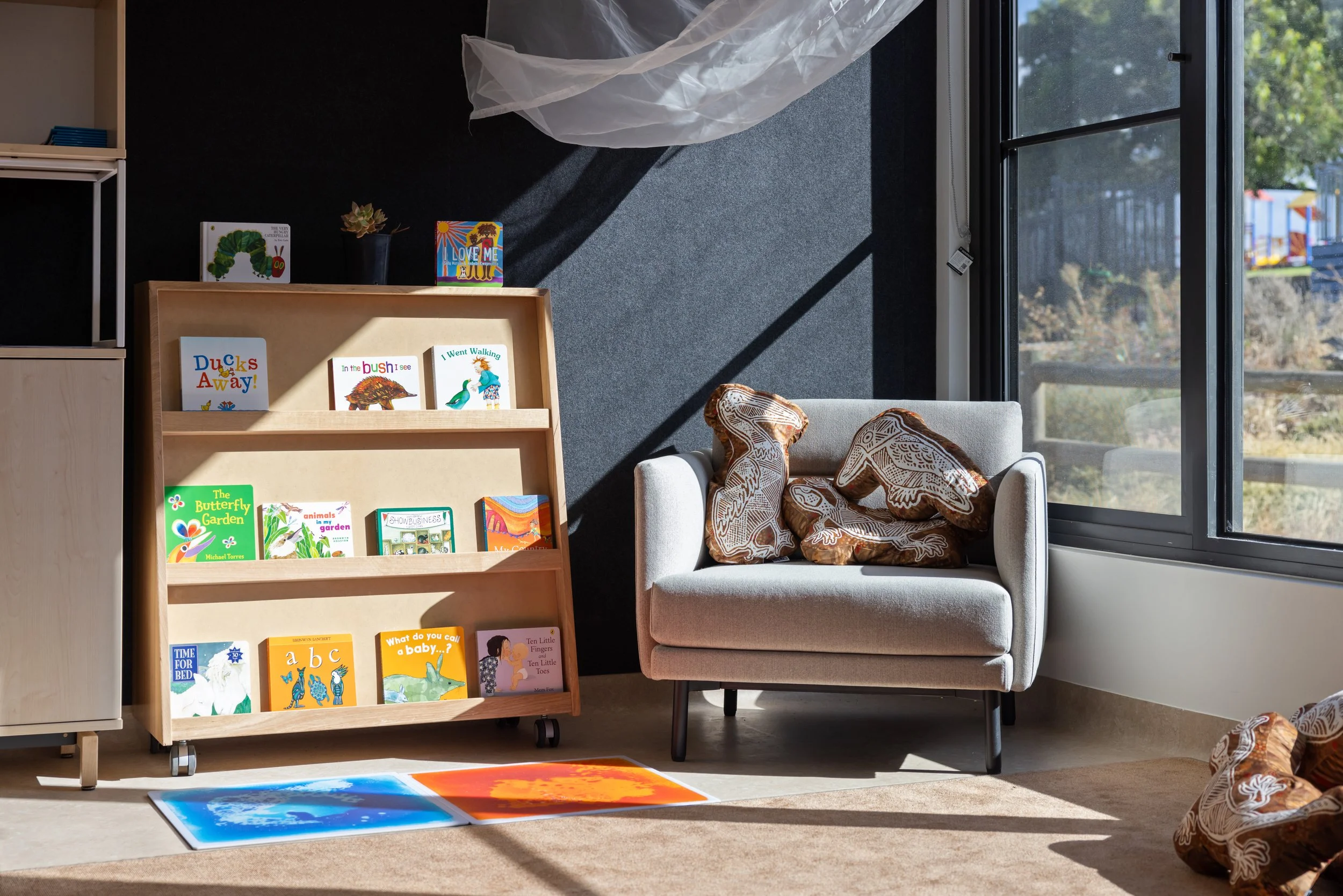 Bright room with a white armchair filled with patterned pillows, a bookshelf on wheels holding children's books, a tan rug on the floor, and large windows letting in natural light, revealing trees and playground outside.