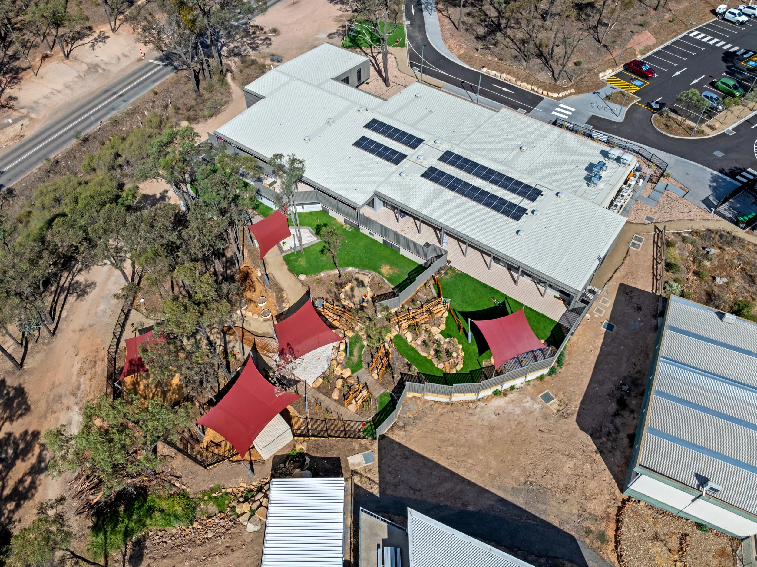 Aerial view of a building with solar panels on its roof, adjacent to a playground with might structures and shaded by trees, in a park-like setting with parking lot nearby.