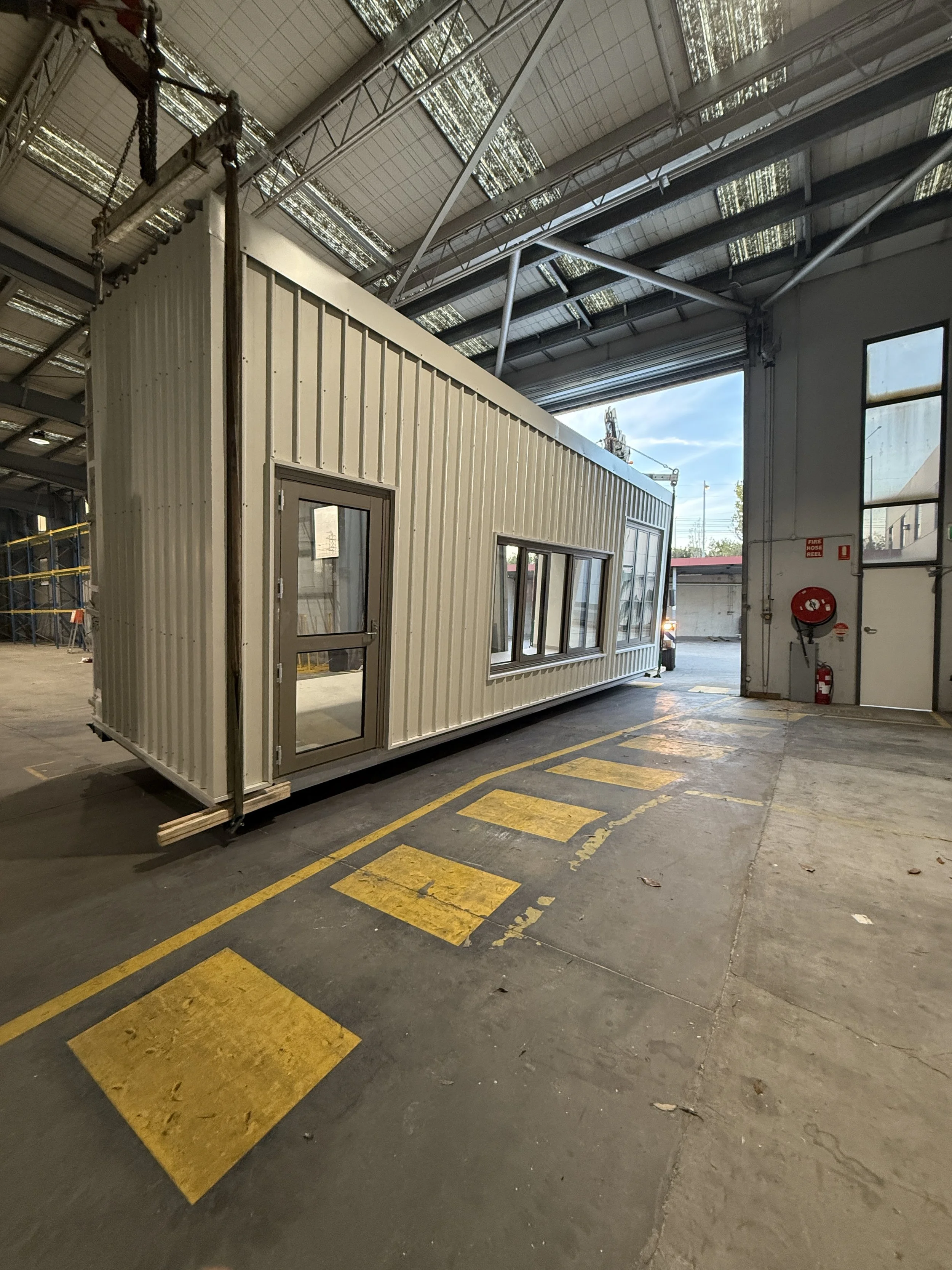 A small, beige modular building with large windows and a door, situated inside an industrial warehouse with a high ceiling and metal framework.