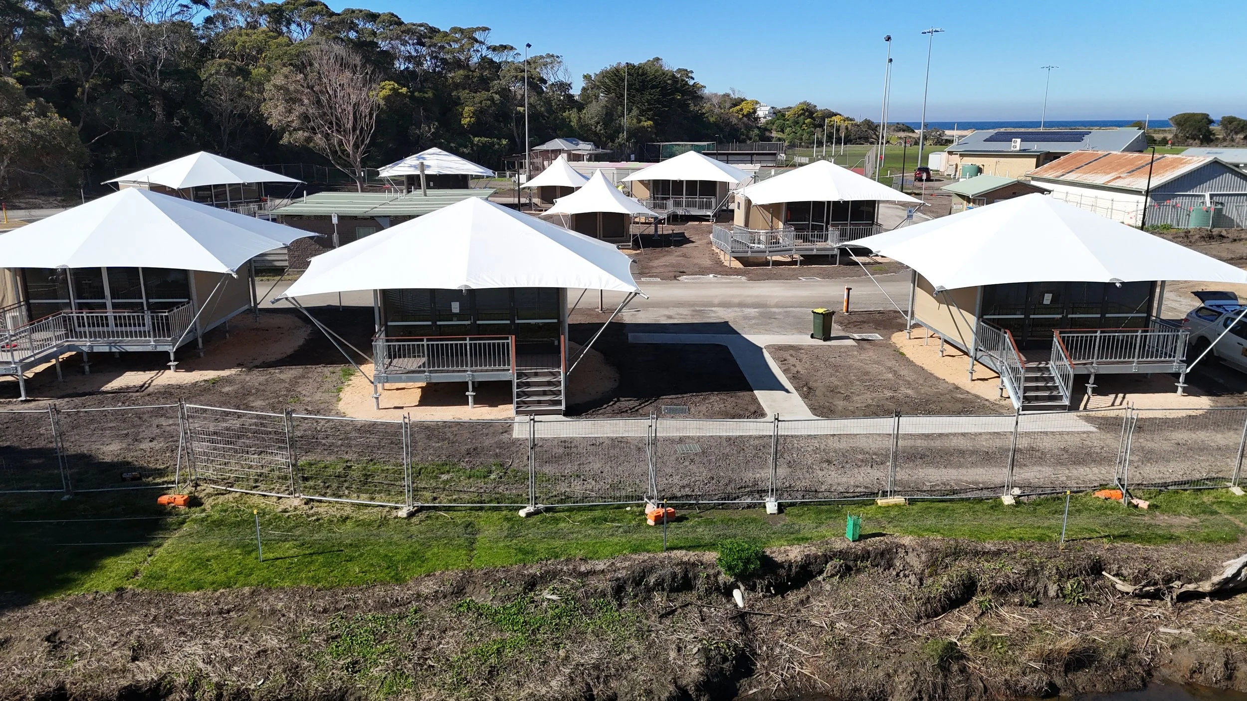 Aerial view of several small buildings with white canopies, located behind a fence with a grassy area in front. The scene appears to be a park or recreational area under construction or renovation, with some dirt and paved pathways visible.