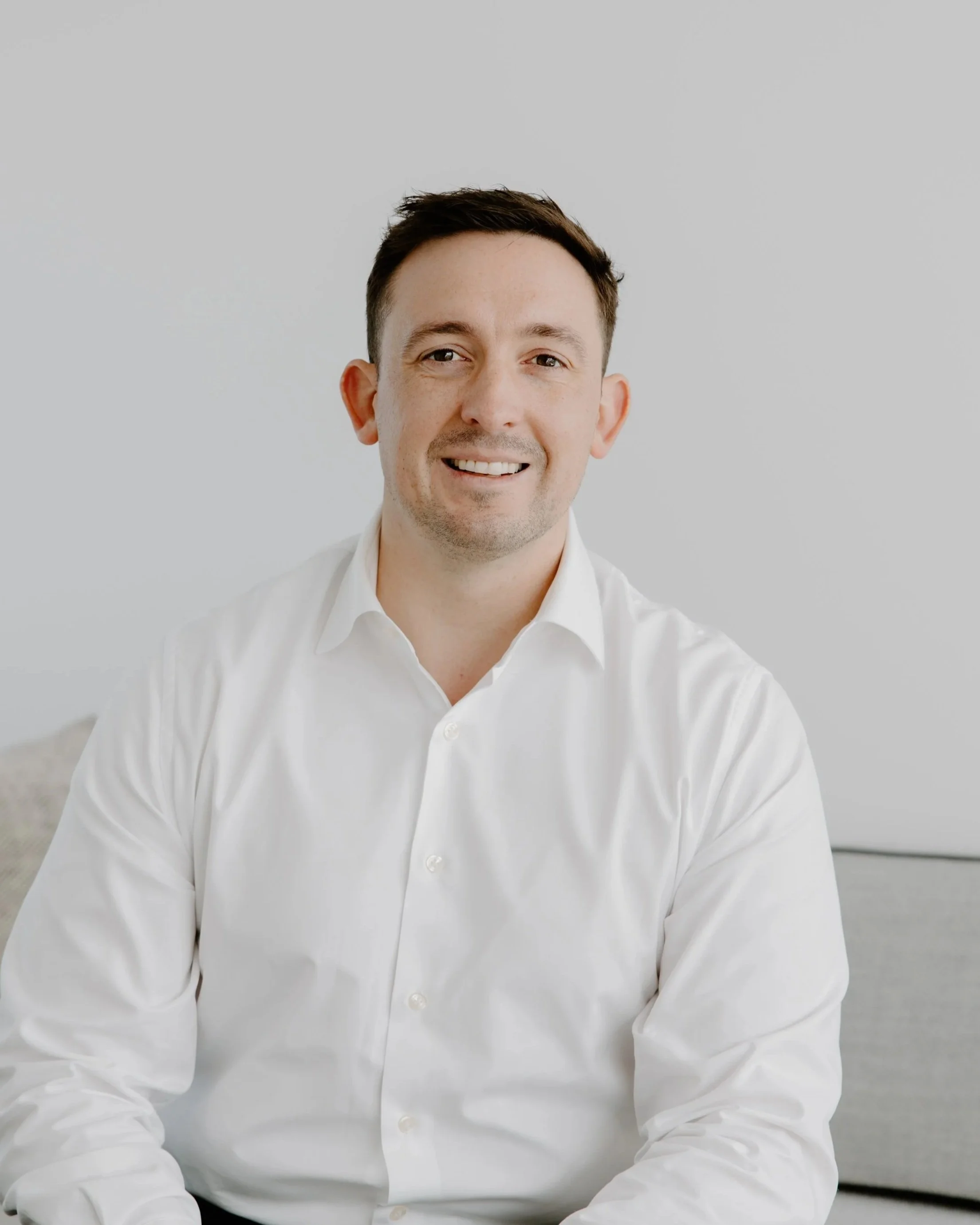 A smiling man in a white shirt sitting against a plain light gray background.