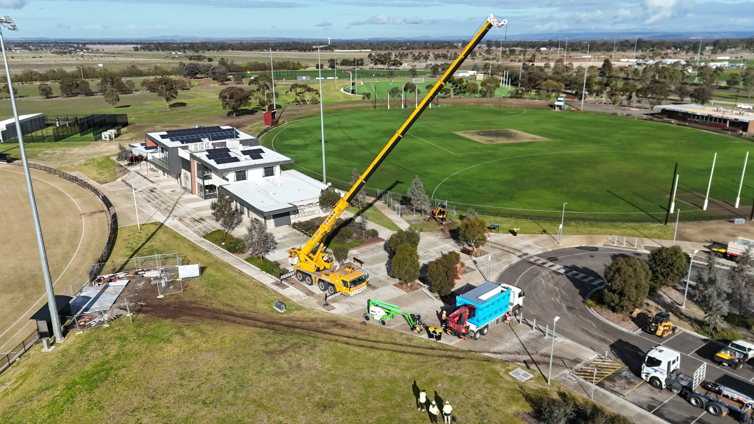 An aerial view of a sports complex with a large yellow crane working near a modern building, and a baseball or cricket field with light poles and other sports fields in the background.