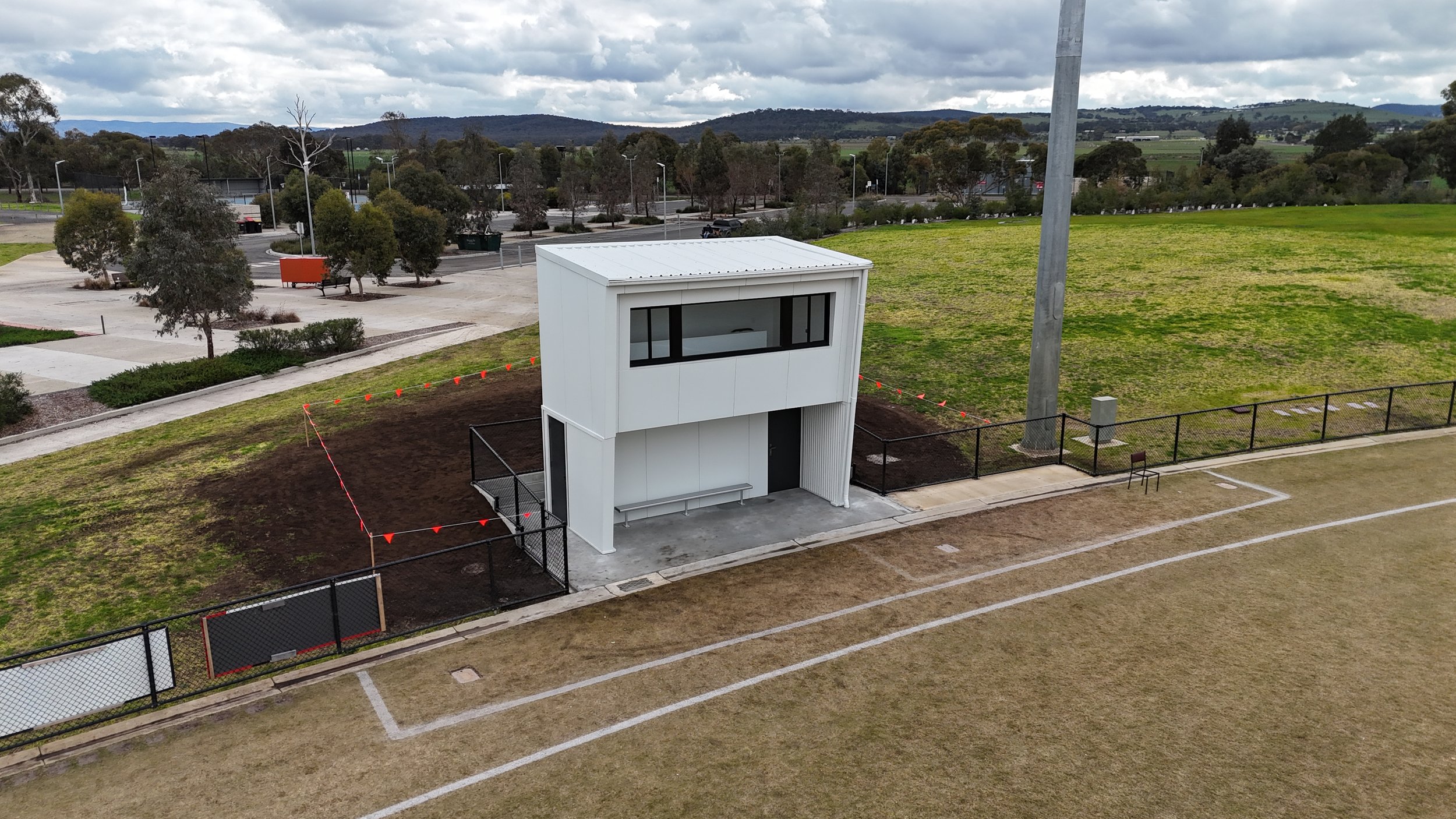 A small modern building with large windows, surrounded by a fenced sports field and bike path, with trees and hills in the background under cloudy skies.