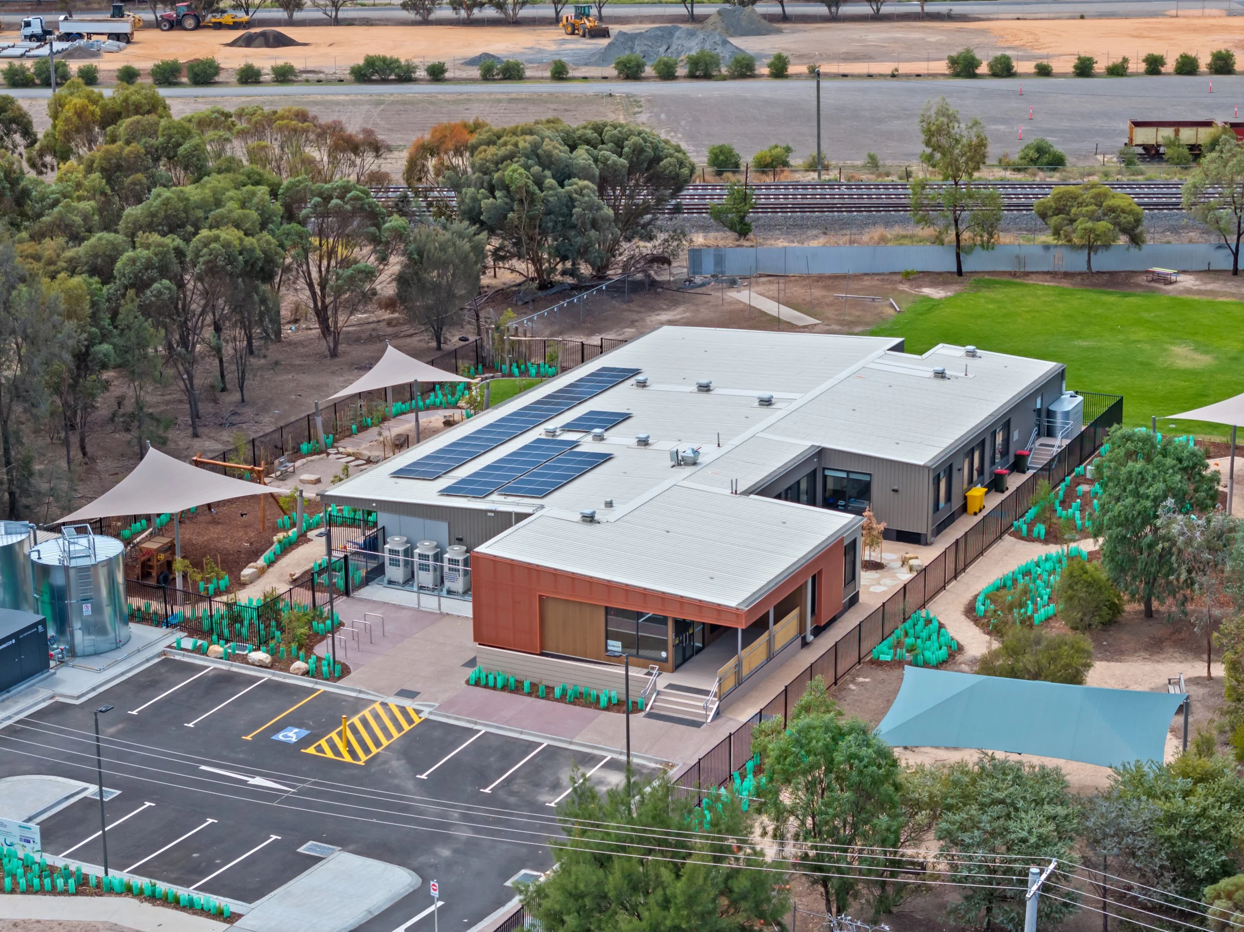 Aerial view of a building with solar panels, a fenced-in yard, landscaped with plants, and a parking lot with two handicapped spots, surrounded by trees and a construction site in the background.