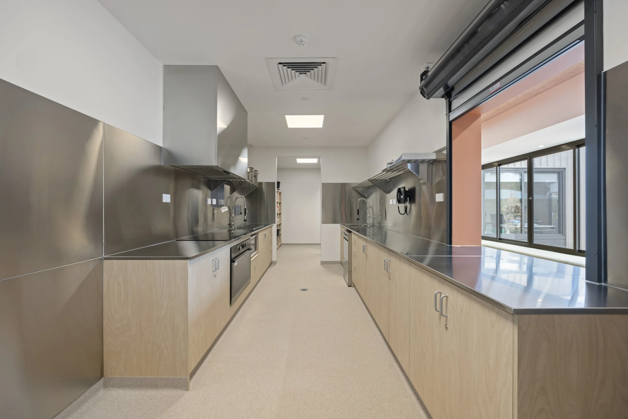 A clean, empty commercial kitchen with stainless steel countertops, wooden cabinets, and large windows.