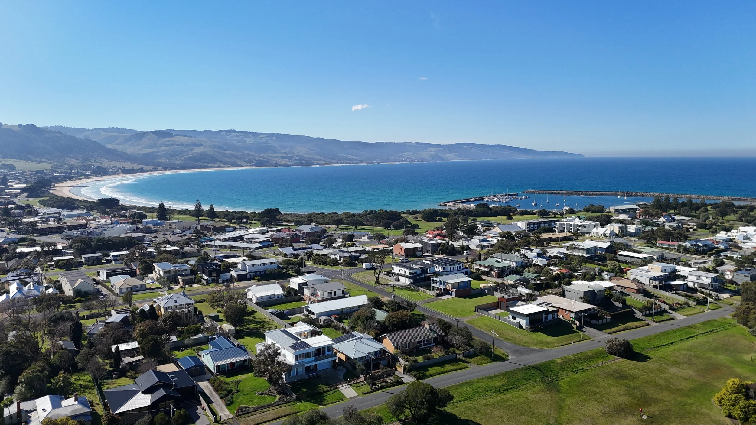 Aerial view of a coastal town with residential houses leading to a beach and a pier, with the ocean and hills in the background on a clear, sunny day.