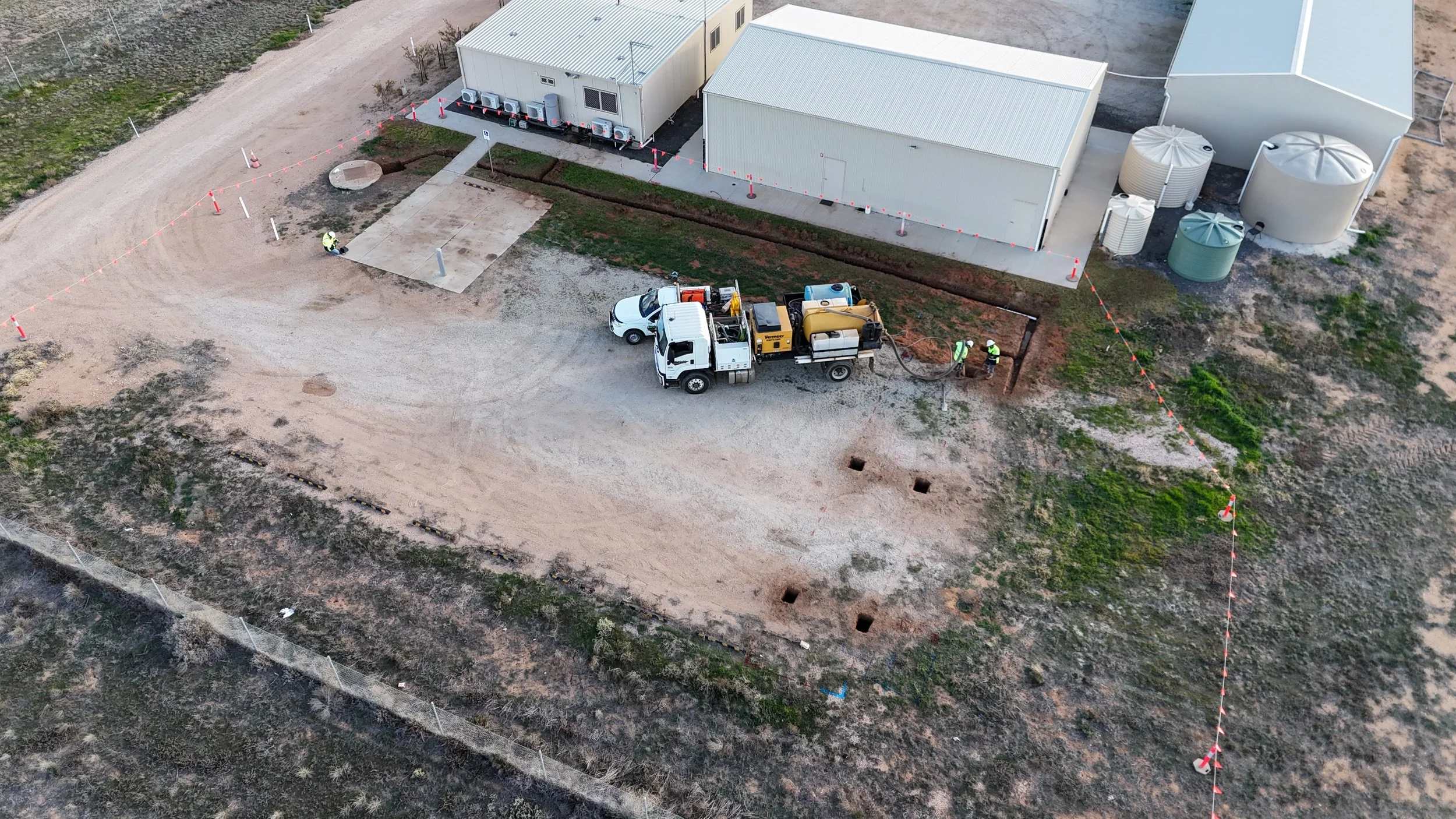 Construction site with dirt road, construction vehicles, workers, and utility buildings, surrounded by orange safety barrier tape.