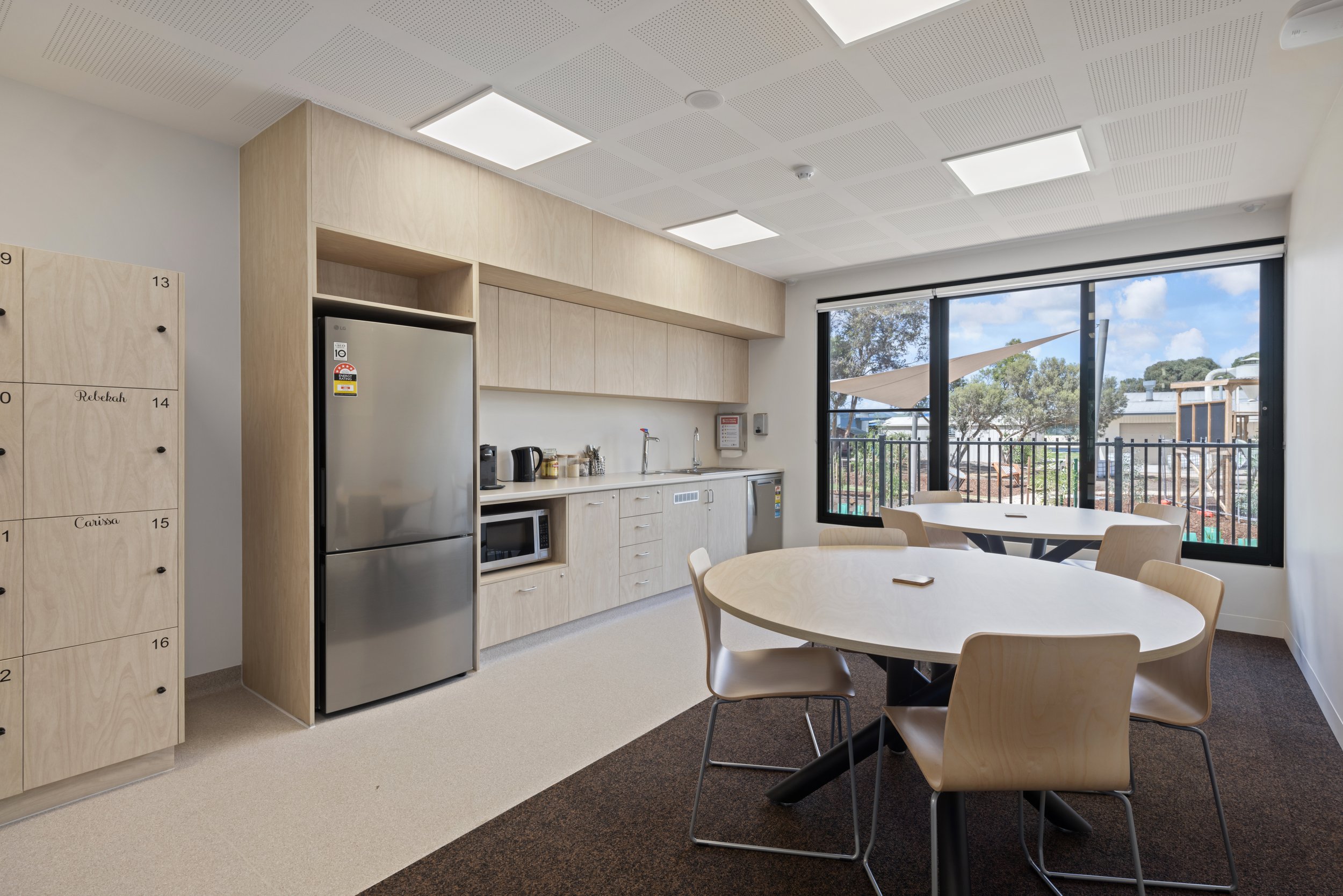 Modern break room with round table, chairs, kitchenette with refrigerator, microwave, coffee maker, and large window overlooking outside patio with trees and blue sky.