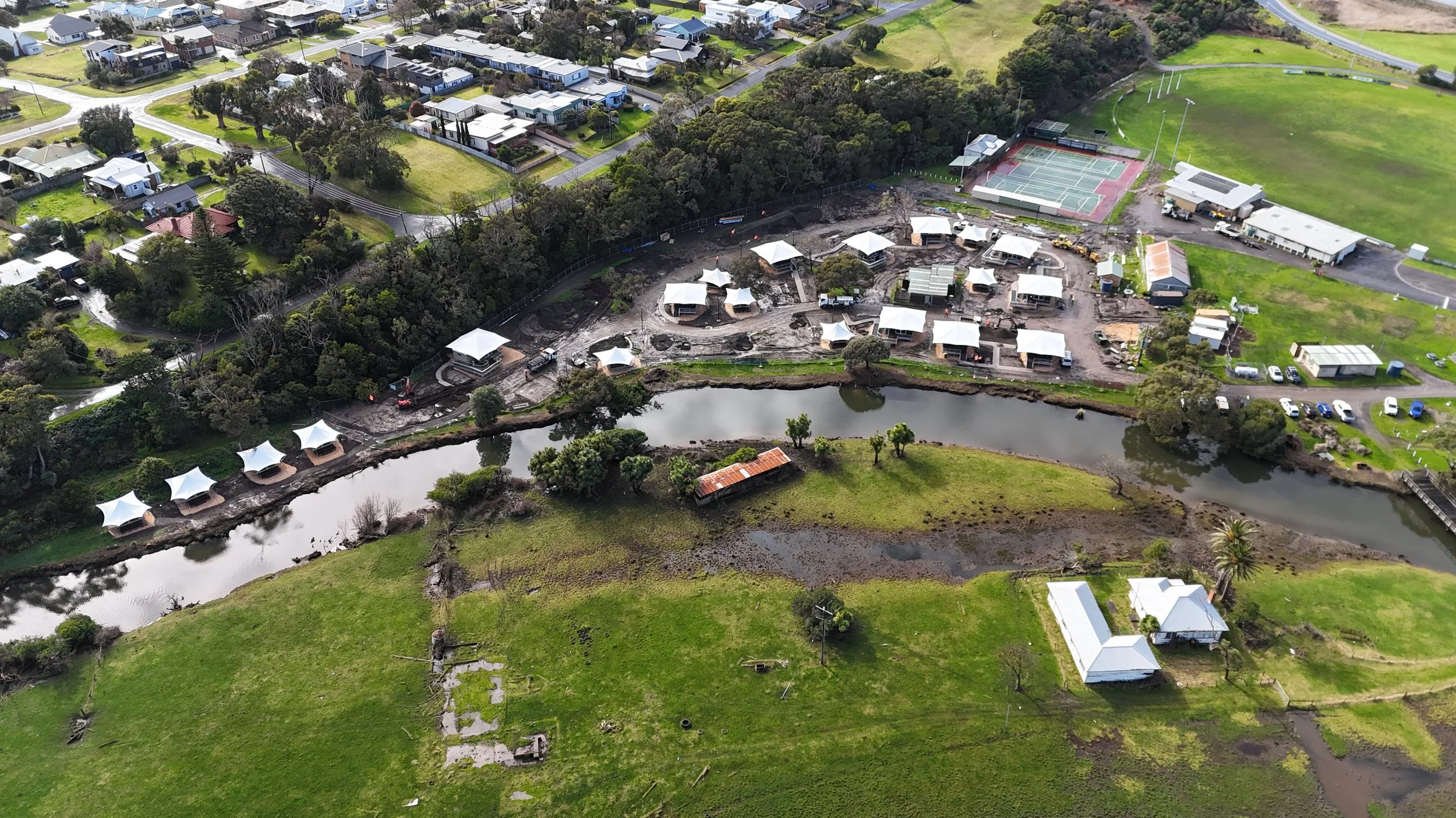 Aerial view of a construction site near a small body of water, with several white temporary structures, nearby houses, a tennis court, and green fields.