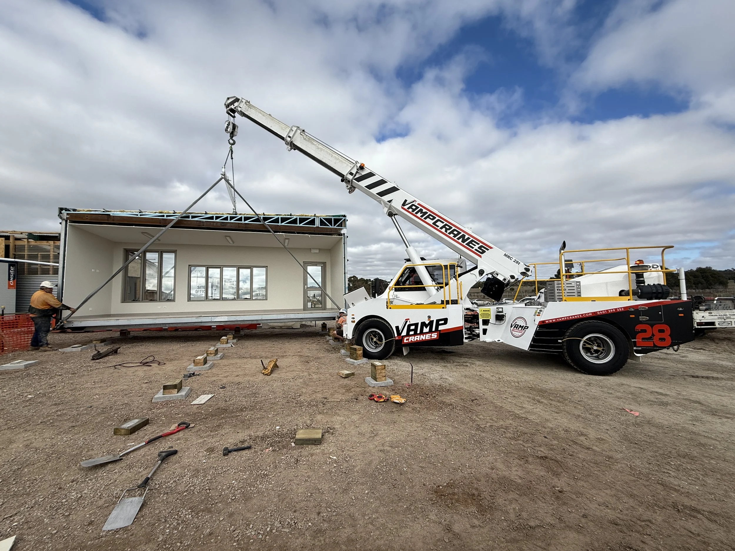 Construction site with a crane lifting a prefabricated building section. Workers are present, and the area is unpaved with construction materials and equipment scattered around.