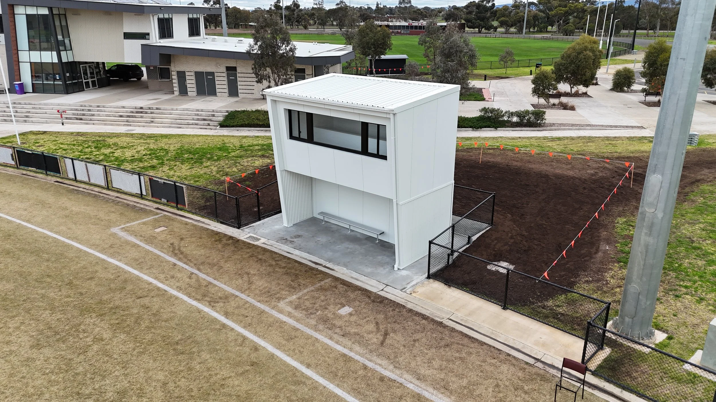 Small modern two-story building with white exterior, large window on upper level, on a sports field with fenced area, nearby athletic facilities, and trees.