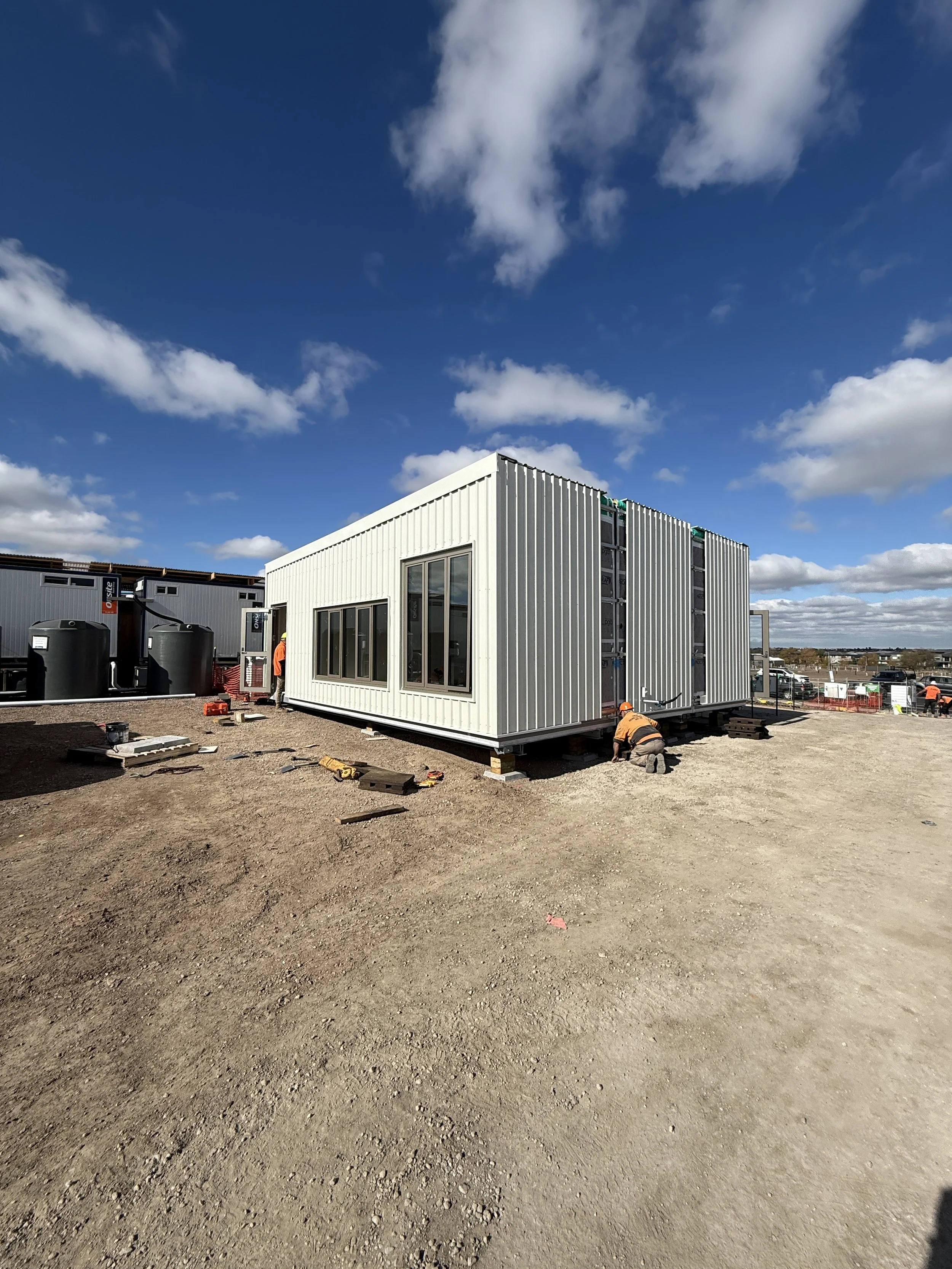 Construction site with a white modular building being assembled, workers in orange safety vests working near it, a clear blue sky with scattered clouds.
