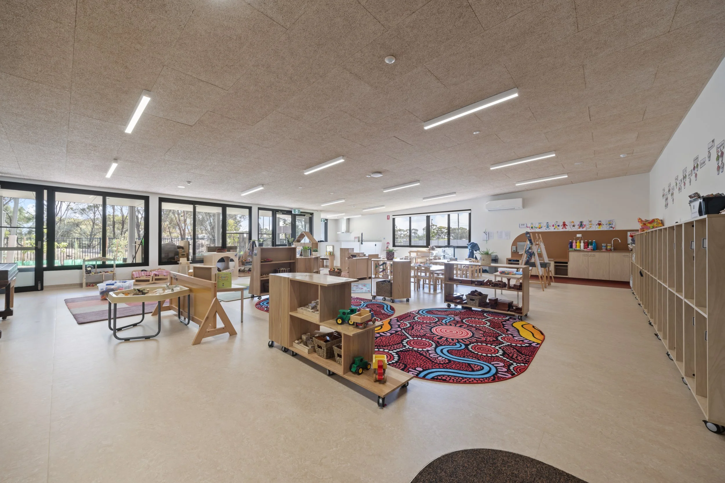 An indoor classroom or playroom with large windows, light wood furniture, toys, and a colorful rug, designed for young children or preschool activities.