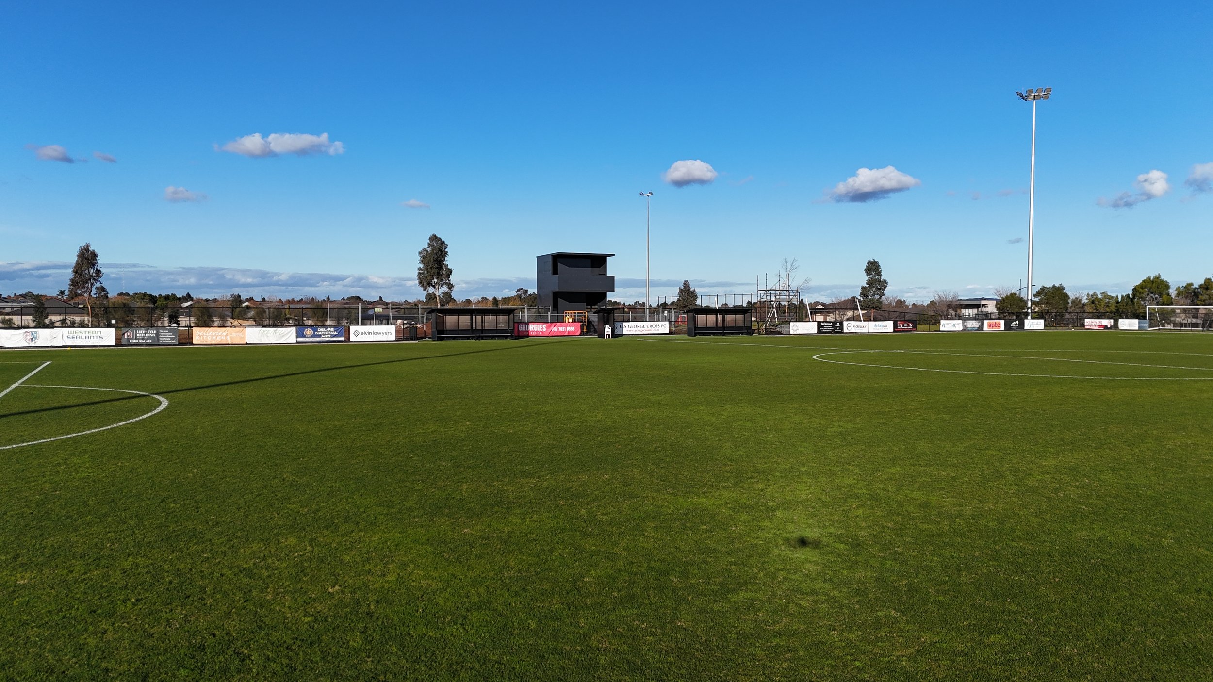 Empty soccer field with green grass, white lines, floodlights, black seating and advertising boards, trees, and a blue sky with some clouds.