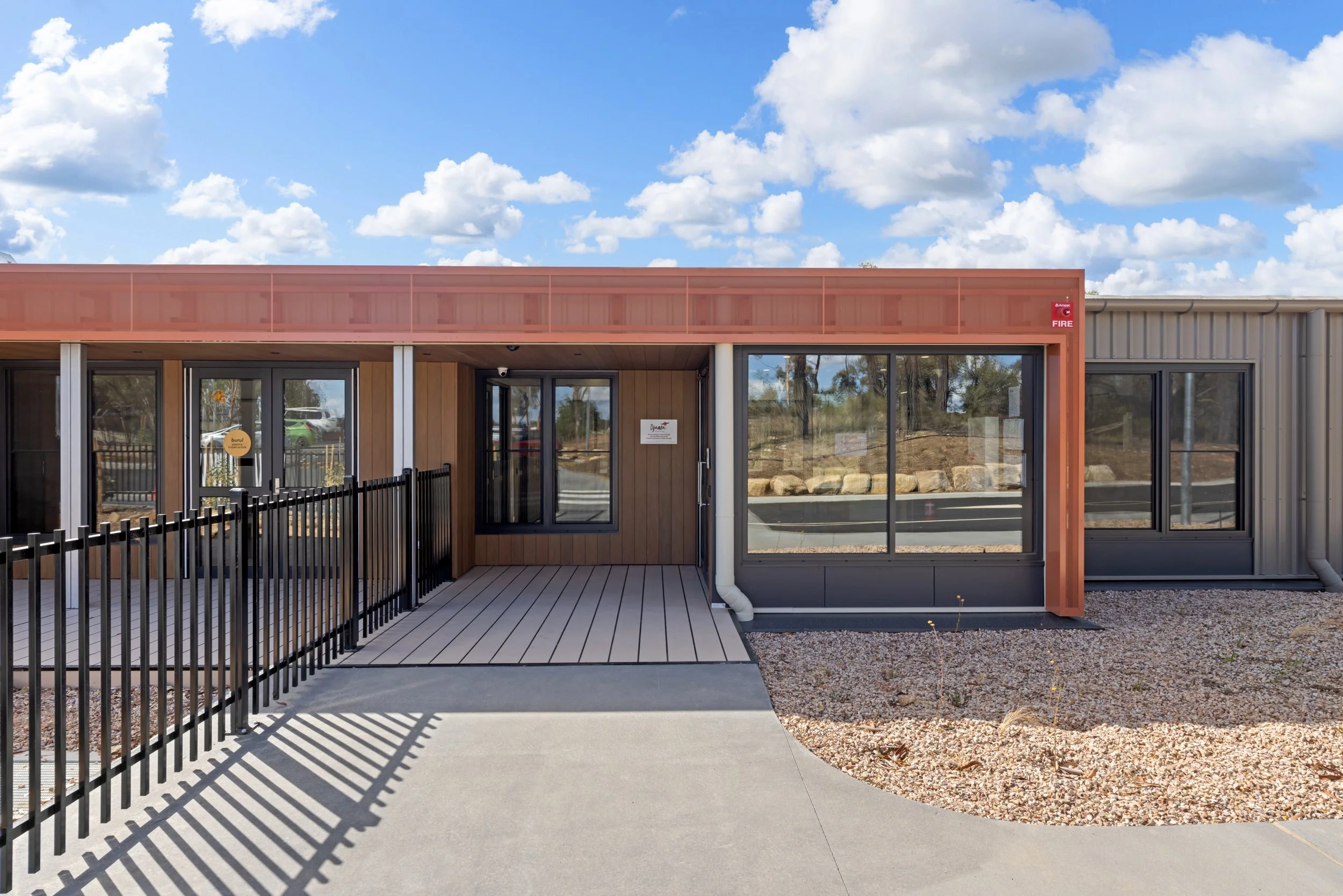 Modern commercial building with wood and metal exterior, black glass doors, and a small fenced patio area, under a partly cloudy sky.