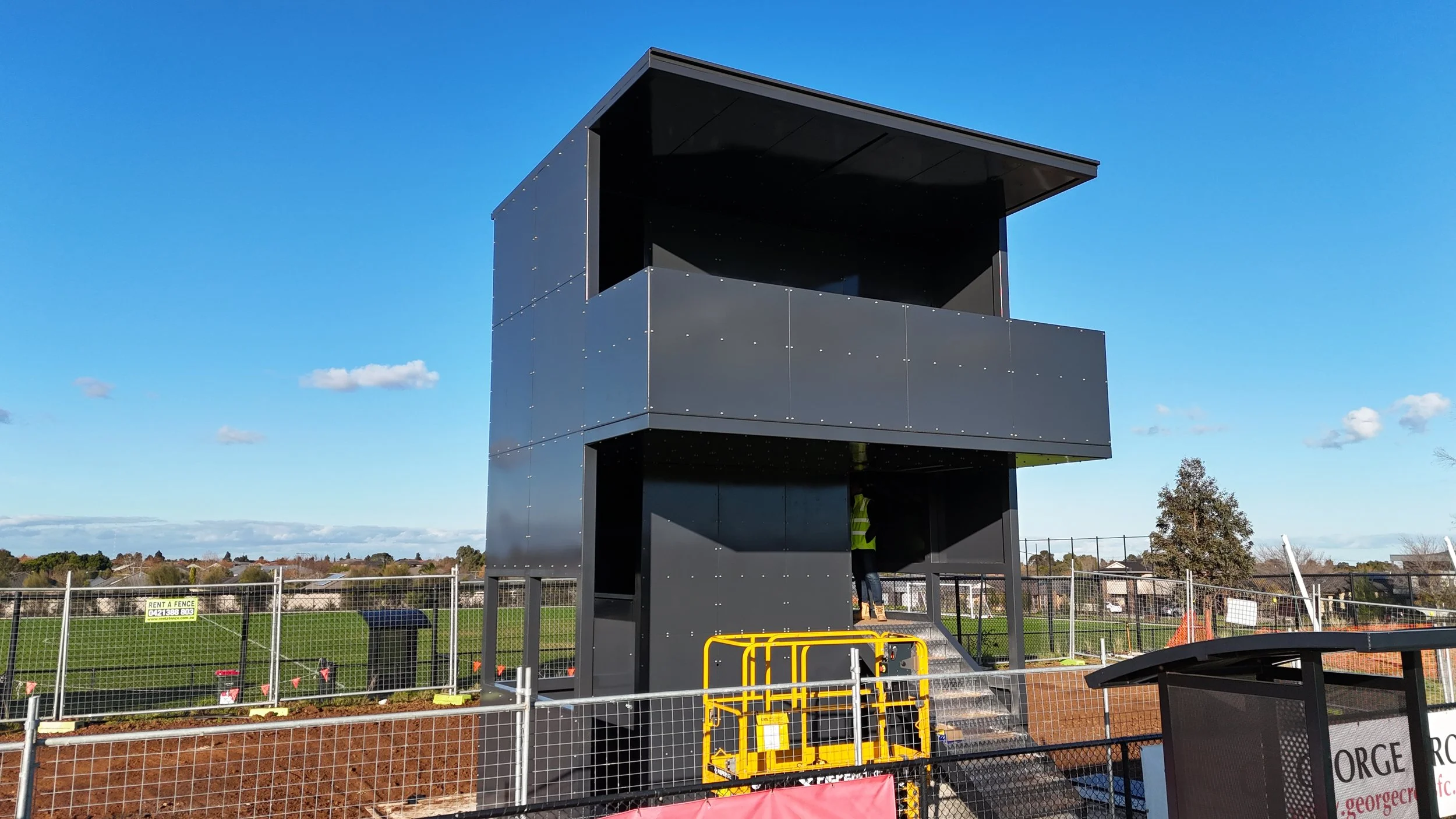 A modern black construction structure with three levels, featuring an open upper level with a roof, situated on a construction site with fencing around it, under a clear blue sky.