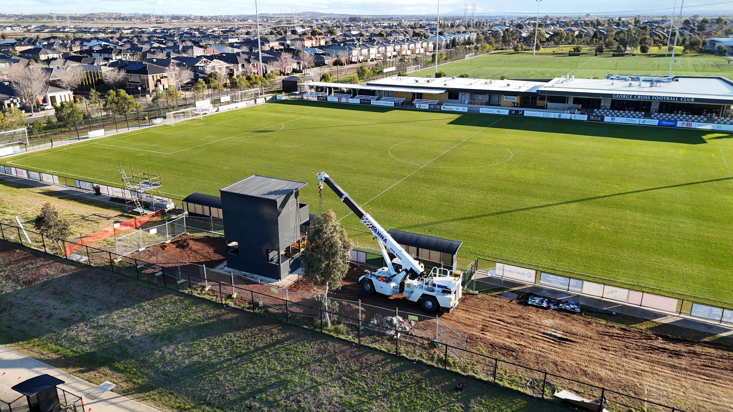 An aerial view of a football stadium under renovation, with a crane installing or repairing equipment near the sideline. The field is well-maintained, surrounded by a fence, with houses and trees beyond the stadium.