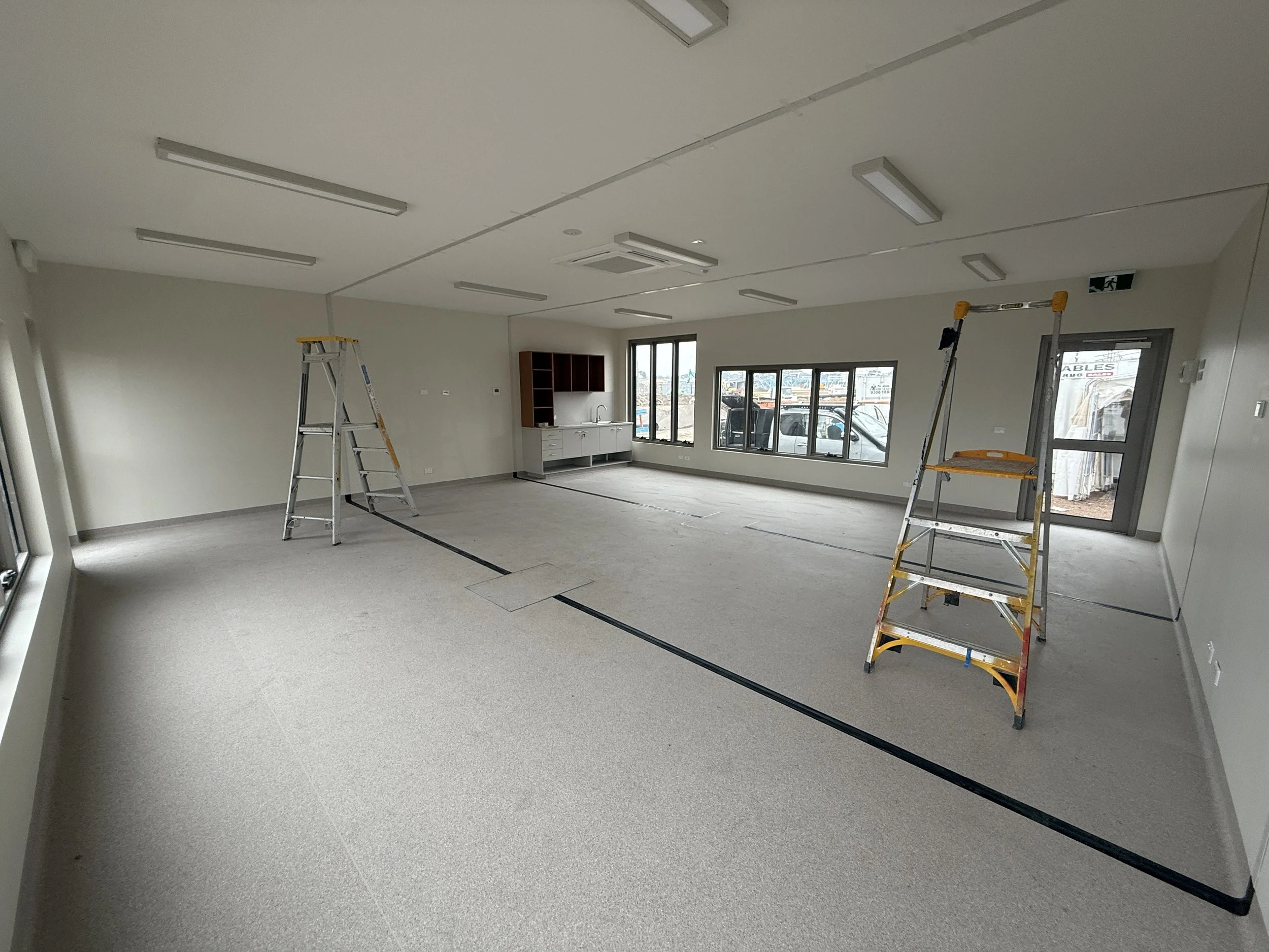 Empty commercial space under renovation with two ladders, a small kitchenette, large windows, and a door leading outside.