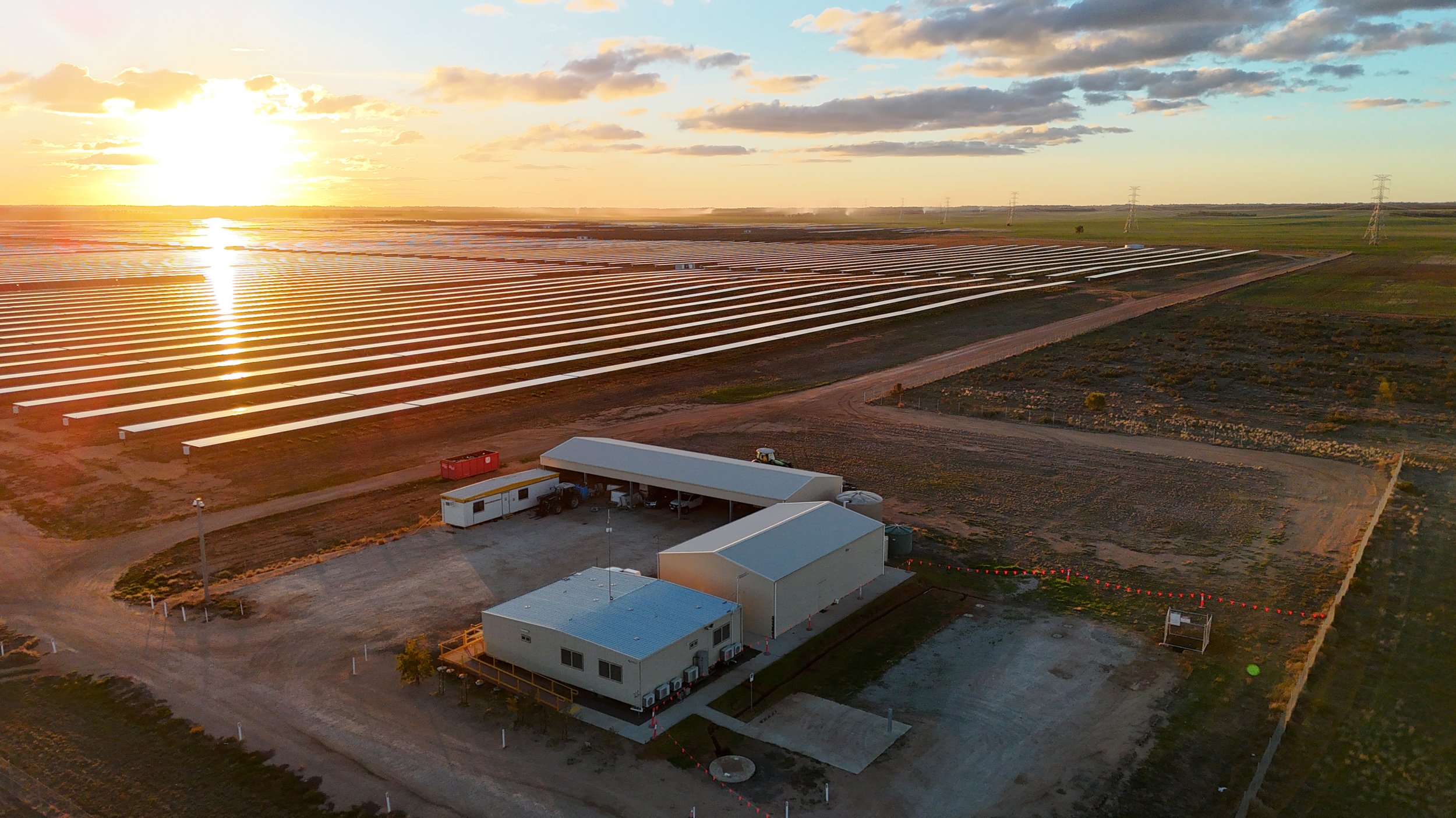 Aerial view of a solar farm with multiple solar panels stretching across a flat landscape, with a few buildings in the foreground, under a partly cloudy sky during sunset.