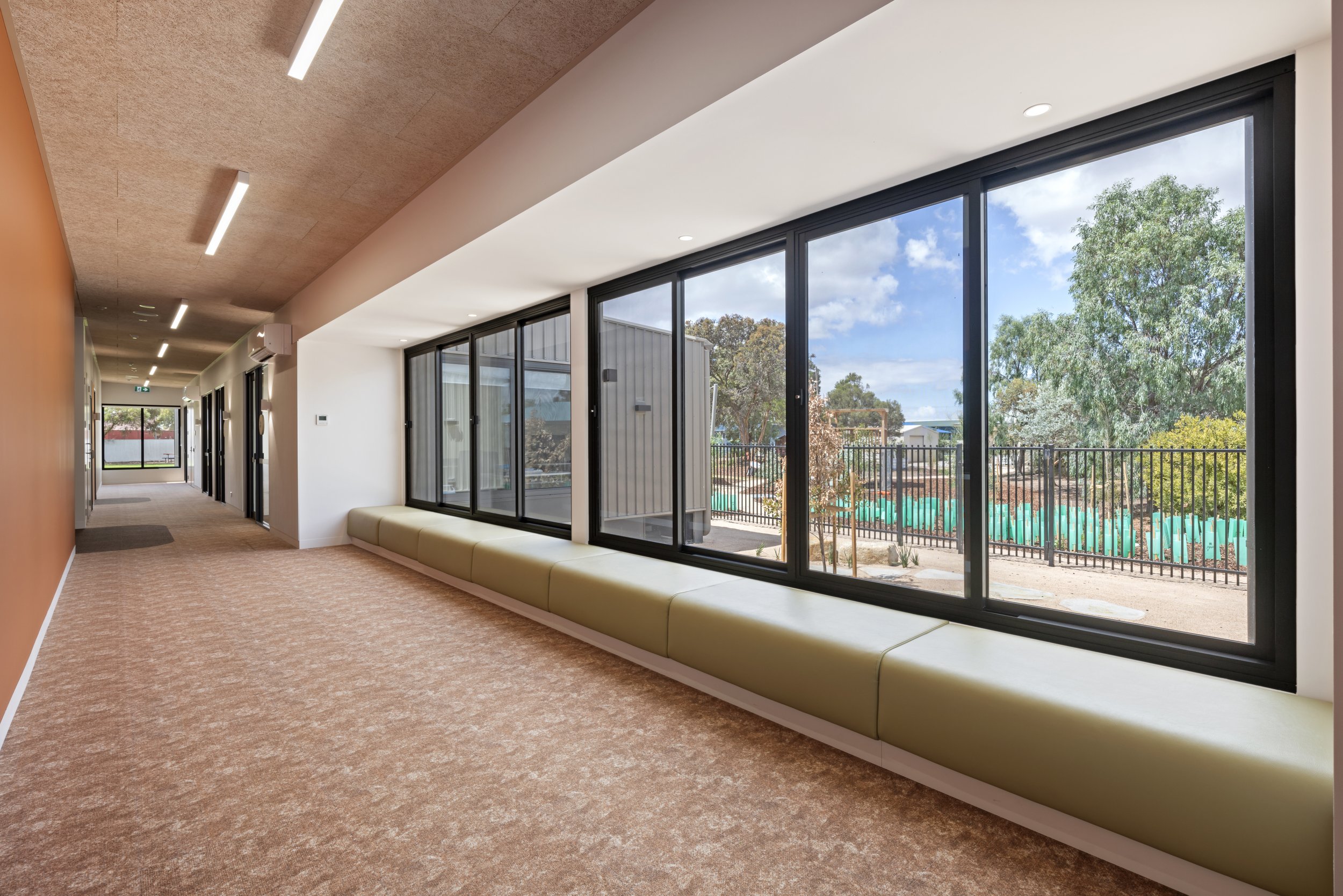 Indoor hallway with large windows showing trees and sky outside. Carpeted floor, beige and white walls, ceiling with recessed and linear light fixtures, and a built-in cushioned seating area along the window.