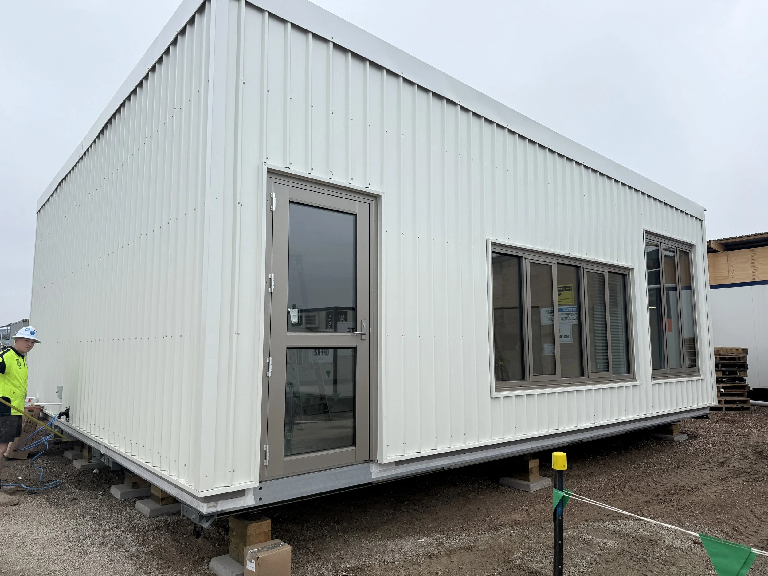 A small white modular building with metal siding and large windows is placed on wooden blocks at a construction site. There is construction equipment and workers visible around.