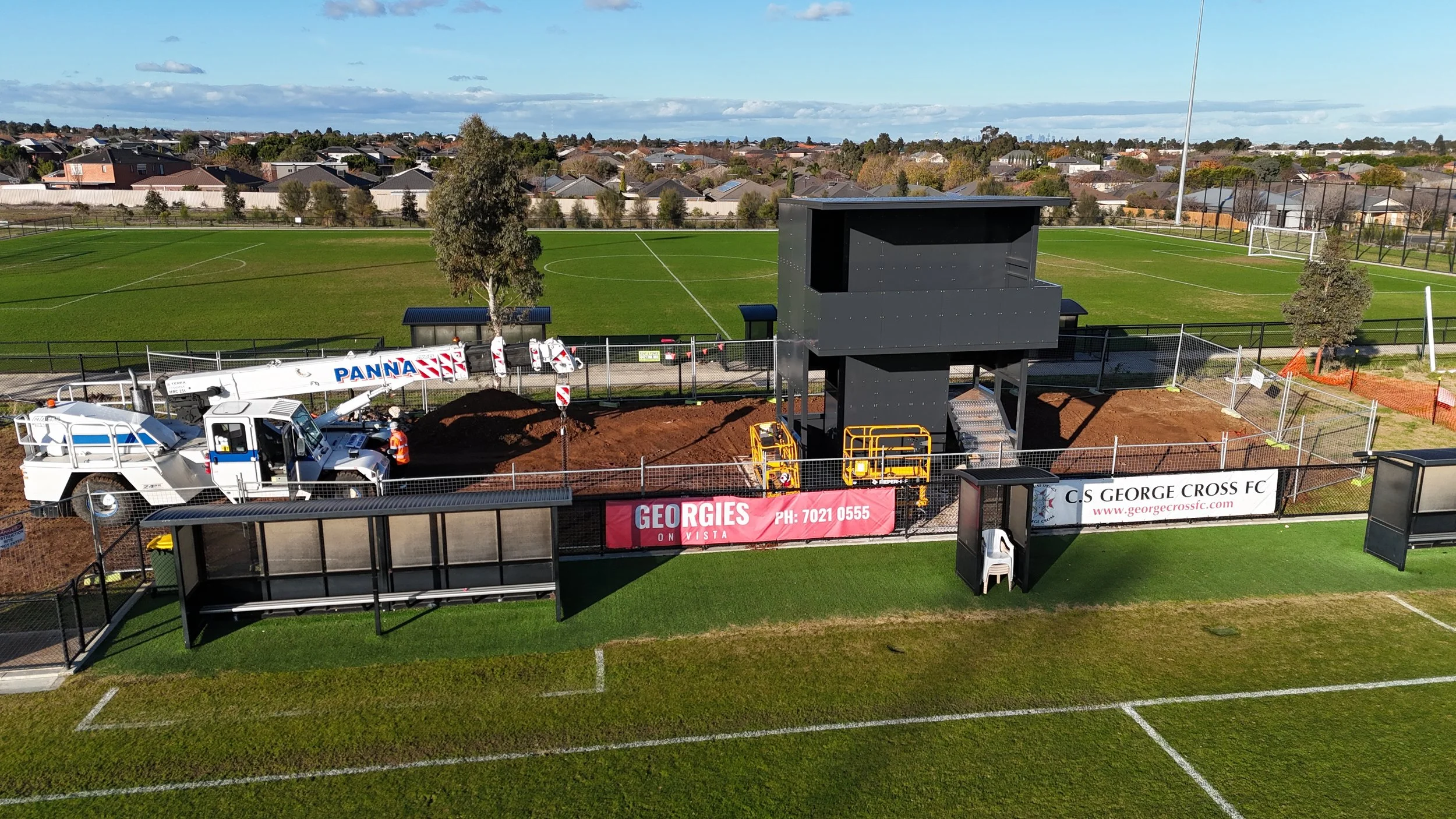 Construction work on a soccer stadium sidelines. Construction vehicle and equipment near a fenced area, with ads and a small shelter. Soccer field visible in the background.